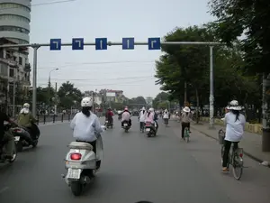Bối cảnh là một con đường lớn, có nhiều làn xe tại một khu đô thị, có thể là Hà Nội. Rất đông người đang tham gia giao thông bằng xe máy và xe đạp. Phía trên đường có một dàn biển báo giao thông màu xanh lam chỉ hướng đi cho các làn xe: hai làn rẽ trái, hai làn đi thẳng và một làn rẽ phải. Nhiều người đi xe máy đội mũ bảo hiểm. Một số người, chủ yếu là phụ nữ, mặc áo khoác chống nắng và đội nón để tránh nắng. Ở giữa bức ảnh là một người phụ nữ đang lái một chiếc xe tay ga màu trắng. Bên trái đường là các tòa nhà, trong đó có một tòa nhà cao tầng hình trụ hiện đại. Bên phải là vỉa hè và hàng cây xanh. Khung cảnh cho thấy một ngày giao thông bình thường ở một thành phố Việt Nam.。（日本語訳）。背景は、ある都市部の、多くの車線がある大通りで、おそらくハノイだろう。非常に多くの人々がバイクや自転車で交通に参加している。道路の上には、各車線の進行方向を示す青い交通標識が一式あり、左折2車線、直進2車線、右折1車線となっている。多くのバイクに乗る人々はヘルメットをかぶっている。一部の人々、主に女性は、日差しを避けるために日焼け防止用のジャケットを着て帽子をかぶっている。写真の中央には、白いスクーターを運転している女性がいる。道路の左側には建物があり、その中には現代的な円筒形の高層ビルが一つある。右側には歩道と緑の並木がある。この光景は、ベトナムのある都市における普通の交通の一日を示している。