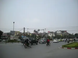 Bức ảnh chụp một ngã tư hoặc bùng binh giao thông đông đúc tại một thành phố ở Việt Nam, có thể là Hà Nội, dưới bầu trời u ám. Nhiều loại phương tiện đang tham gia giao thông trên đường, chủ yếu là xe máy (xe gắn máy) và ô tô. Có thể thấy nhiều người đang điều khiển xe máy, một số chở thêm người ngồi sau, và hầu hết đều đội mũ bảo hiểm. Các loại ô tô bao gồm một chiếc SUV màu đen, một chiếc hatchback màu bạc và các xe sedan khác. Ở phía xa, một dòng xe cộ đang di chuyển, trong đó có cả người đi xe đạp. Hệ thống đèn giao thông đang hoạt động để điều tiết dòng xe, với một số đèn báo màu đỏ và một số báo màu xanh. Xung quanh ngã tư là các tòa nhà nhiều tầng san sát nhau, đặc trưng của đô thị. Phía trên là mạng lưới dây điện chằng chịt. Ở giữa đường có một dải phân cách hoặc vòng xuyến được trồng cỏ và có lề đường sơn hai màu đỏ trắng. Có các biển hiệu trên các tòa nhà nhưng chữ viết không thể đọc rõ.。（日本語訳）。この写真は、曇り空の下、ベトナムのある都市、おそらくハノイの、交通量の多い交差点またはロータリーを撮影したものです。道路では多くの種類の乗り物が交通に参加しており、主にバイク（原付バイク）と自動車です。多くの人々がバイクを運転しているのが見え、何人かは後ろに同乗者を乗せており、ほとんどがヘルメットを着用しています。自動車の種類には、黒いSUV一台、銀色のハッチバック一台、その他のセダンなどが含まれます。遠方では、車両の流れが動いており、その中には自転車に乗る人もいます。交通信号システムが車両の流れを調整するために作動しており、いくつかの信号は赤色を、いくつかは青色を示しています。交差点の周りには、都市に特徴的な、多層階の建物が密集しています。上方には、複雑に絡み合った電線の網があります。道路の中央には、草が植えられた中央分離帯またはロータリーがあり、赤と白の二色で塗られた縁石があります。建物には看板がありますが、文字ははっきりと読み取れません。