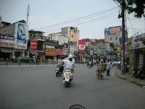 Bối cảnh là một con phố đông đúc ở Hà Nội với nhiều phương tiện giao thông, chủ yếu là xe máy. Mọi người đang điều khiển xe máy trên đường, một số người đội mũ bảo hiểm. Một người đi xe máy đang chở các thùng hàng cồng kềnh được xếp chồng lên nhau ở yên sau. Hai bên đường là những dãy nhà san sát, tầng trệt là các cửa hàng kinh doanh với rất nhiều biển hiệu quảng cáo. Phía trên các tòa nhà có một cột tháp viễn thông và hệ thống dây điện chằng chịt.。Các dòng chữ trên các biển hiệu có thể đọc được bao gồm:。- Phía bên trái: "BẾN NGHÉ", "Thanh Hà", "ĐÚT BÌA - VĂN PHÒNG PHẨM", một biển quảng cáo lớn màu xanh cho "MÁY TRỢ THÍNH" của hãng "SIEMENS" và tên cửa hàng "CÁT TƯỜNG", "PHOTOCOPY", "SIM SỐ ĐẸP", "CẮT TÓC NAM NỮ", và số nhà "134".。- Phía bên phải: "KIPOR", một biển hiệu khác của "BẾN NGHÉ", "MÁY IN - MÁY FAX - MÁY PHOTOCOPY", "VIỆN THÔNG TRẦN...". Ở phía xa, có một biển quảng cáo lớn trên nóc nhà với chữ "NGK" và "G-POWER".。（日本語訳）。背景はハノイの混雑した通りで、多くの交通手段、主にバイクが見られます。人々は道路でバイクを運転しており、何人かはヘルメットを着用しています。あるバイクの運転手は、後部座席に積み重ねられたかさばる荷物を運んでいます。道路の両側には家々が密集しており、1階は多くの広告看板を掲げた店舗になっています。建物の​​上には通信塔と、複雑に絡み合った電線があります。看板に書かれている読み取れる文字は以下の通りです：。- 左側：「BẾN NGHÉ」、「Thanh Hà」、「ĐÚT BÌA - VĂN PHÒNG PHẨM（製本 - 文房具）」、「SIEMENS」社の「MÁY TRỢ THÍNH（補聴器）」の大きな青い広告看板と店名「CÁT TƯỜNG」、「PHOTOCOPY（コピー）」、「SIM SỐ ĐẸP（美しい番号のSIM）」、「CẮT TÓC NAM NỮ（男女ヘアカット）」、そして番地の「134」。- 右側：「KIPOR」、「BẾN NGHÉ」の別の看板、「MÁY IN - MÁY FAX - MÁY PHOTOCOPY（プリンター - ファックス - コピー機）」、「VIỆN THÔNG TRẦN...（チャン通信...）」。遠くには、屋上に「NGK」と「G-POWER」の文字が入った大きな広告看板があります。