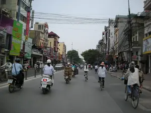 Bức ảnh chụp một con phố đông đúc và nhộn nhịp ở một thành phố Việt Nam, có thể là Hà Nội. Dòng người và phương tiện di chuyển tấp nập trên đường, chủ yếu là xe máy (xe số và xe tay ga) và xe đạp. Một số người đi xe máy đội mũ bảo hiểm, trong khi những người đi xe đạp thì đội mũ. Một người đi xe máy đang chở một kiện hàng lớn được buộc ở phía sau. Hai bên đường là những dãy nhà phố cao tầng, san sát nhau, một kiến trúc đặc trưng của các đô thị Việt Nam. Phía trên là một hệ thống dây điện và dây cáp chằng chịt. Có rất nhiều biển hiệu quảng cáo trên các tòa nhà. Một số biển hiệu có thể đọc được là: 'SIÊU THỊ', 'NỘI THẤT GIA ĐÌNH VÀNG', 'HOTEL', 'Silk LỤA VIỆT', 'ÁO DÀI', 'Achi', 'VÍ DA'. Những biển hiệu này cho thấy đây là một khu phố thương mại với nhiều loại hình kinh doanh.。（日本語訳）。この写真はベトナムの都市、おそらくハノイの、混雑して賑やかな通りを撮影したものです。道路上では人々と乗り物がひっきりなしに行き交っており、主にバイク（マニュアル車とスクーター）と自転車です。バイクに乗っている人の中にはヘルメットを着用している人もいれば、自転車に乗っている人は帽子をかぶっています。バイクに乗っている一人が、後ろに縛り付けられた大きな荷物を運んでいます。通りの両側には、ベトナムの都市に特有の建築様式である、高層のタウンハウスが密集して立ち並んでいます。頭上には、電線やケーブルが複雑に絡み合ったシステムがあります。建物にはたくさんの広告看板があります。読み取れる看板には、「SIÊU THỊ（スーパーマーケット）」、「NỘI THẤT GIA ĐÌNH VÀNG（ゴールデンファミリーインテリア）」、「HOTEL（ホテル）」、「Silk LỤA VIỆT（ベトナムシルク）」、「ÁO DÀI（アオザイ）」、「Achi」、「VÍ DA（革財布）」などがあります。これらの看板は、ここが様々な業種の商業地区であることを示しています。