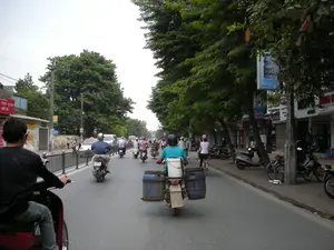 Đây là một cảnh đường phố ở Việt Nam, có thể là Hà Nội, với mật độ giao thông đông đúc, chủ yếu là xe máy. Mọi người đang di chuyển trên đường, hầu hết đều đội mũ bảo hiểm. Hai bên đường là những hàng cây xanh và các tòa nhà có cửa hàng ở tầng trệt. Một người đàn ông đang lái một chiếc xe máy được sửa đổi để chở hàng, với hai thùng phuy lớn màu xanh và một thùng xốp trắng được buộc chặt phía sau. Những người khác cũng đang đi xe máy, và có một người phụ nữ đang đi xe đạp. Trên vỉa hè bên phải, có nhiều xe máy đang đậu trước các cửa hàng. Có một số biển hiệu có thể nhận dạng được. Ở bên trái, một tấm biển màu đỏ ghi: 'SỬA ĐỒNG HỒ - CẮT KÍNH - ĐÁ HAY - CẤP SỐ' và một biển hiệu khác ghi 'PHOTOCOPY'. Treo trên một cái cây là một biển quảng cáo cho sản phẩm sữa uống lên men 'Probi'. Các biển hiệu khác của các cửa hàng ở phía xa hơn thì không thể đọc rõ.。（日本語訳）。これはベトナム、おそらくハノイの街頭風景で、交通量が多く、主にバイクです。人々は道路を移動しており、ほとんどがヘルメットを着用しています。道路の両側には緑の並木と、1階に店舗が入った建物があります。一人の男性が、荷物を運ぶために改造されたバイクを運転しており、後ろには2つの大きな青いドラム缶と1つの白い発泡スチロールの箱がしっかりと結び付けられています。他の人々もバイクに乗っており、一人の女性が自転車に乗っています。右側の歩道には、店の前に多くのバイクが駐車されています。いくつか識別できる看板があります。左側には、赤い看板に「時計修理 - ガラスカット - ライター修理 - 番号発行」と書かれており、別の看板には「PHOTOCOPY」と書かれています。木には、乳酸菌飲料「Probi」の広告看板が掛かっています。さらに遠くにある店の他の看板は、はっきりと読み取れません。