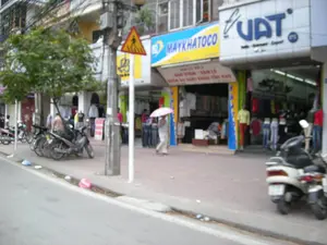 Đây là hình ảnh một con phố ở thành thị với các cửa hàng kinh doanh san sát nhau. Nổi bật là hai cửa hàng quần áo. Một cửa hàng có biển hiệu màu xanh và vàng ghi chữ 'MAYKHATOCO'. Bên dưới biển hiệu này có các dòng chữ nhỏ hơn: 'BÁN LẺ SỐ 2', 'BÁN BUÔN - BÁN LẺ', 'NGUỒN LÔ XUẤT KHẨU TỒN KHO'. Cửa hàng bên cạnh có biển hiệu lớn màu trắng với chữ 'UAT' màu xanh dương và dòng chữ 'Textile - Garment - Export'. Các cửa hàng đang mở cửa, trưng bày quần áo trên các ma-nơ-canh và giá treo. Trên vỉa hè lát gạch, một người đang đi bộ và che một chiếc ô màu hồng. Có nhiều xe máy đang đậu dọc theo vỉa hè. Phía trước có một cột điện và một biển báo giao thông hình tam giác nền vàng, viền đỏ với biểu tượng người đi bộ, cảnh báo khu vực dành cho người đi bộ qua đường.。（日本語訳）。これは、商店が隣接する都市の通りの画像です。目立つのは2軒の衣料品店です。一つの店には「MAYKHATOCO」と書かれた青と黄色の看板があります。この看板の下には、「BÁN LẺ SỐ 2」、「BÁN BUÔN - BÁN LẺ」、「NGUỒN LÔ XUẤT KHẨU TỒN KHO」という、より小さな文字の行があります。隣の店には、青い文字で「UAT」と「Textile - Garment - Export」という文字が書かれた大きな白い看板があります。店は開店しており、マネキンやハンガーラックに服を陳列しています。タイル敷きの歩道の上を、一人の人がピンク色の傘をさして歩いています。歩道に沿ってたくさんのバイクが駐車しています。前方には電柱が1本と、黄色い背景に赤い縁取り、歩行者のシンボルが描かれた三角形の交通標識があり、歩行者横断区域であることを警告しています。