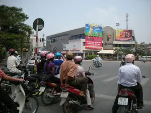Bức ảnh ghi lại cảnh giao thông tại một ngã tư đường phố đông đúc ở Hà Nội vào ban ngày. Rất nhiều người đang điều khiển xe máy và dừng lại để chờ tín hiệu đèn giao thông. Tất cả những người đi xe máy đều đội mũ bảo hiểm với nhiều màu sắc khác nhau như hồng, xanh dương, đỏ và trắng.。Ở phía sau là một tòa nhà lớn, là trung tâm thương mại Pico Plaza. Trên tòa nhà có nhiều biển quảng cáo lớn:。- Một biển quảng cáo màu đỏ nổi bật của Pico Plaza với dòng chữ: "PICO PLAZA", "HỆ THỐNG SIÊU THỊ ĐIỆN MÁY", "HÀNG ĐẦU VIỆT NAM".。- Một biển quảng cáo khác dành cho nước giải khát C2 với dòng chữ "mát lạnh & thanh khiết".。- Có thể thấy logo của "S-Fone" trên tòa nhà.。Các chi tiết khác trong ảnh bao gồm một cột đèn giao thông đang bật đèn xanh, một cột biển báo giao thông hình tròn màu xanh lá cây có chữ viết nhưng khó đọc rõ, cây xanh bên lề đường, và một người đang đi xe đạp. Khung cảnh này đặc trưng cho sự nhộn nhịp của giao thông đô thị tại Việt Nam.。（日本語訳）。この写真は、ハノイの昼間の混雑した交差点の交通風景を記録したものです。非常に多くの人々がバイクを運転し、信号を待つために停止しています。バイクに乗っている人々は皆、ピンク、青、赤、白など、さまざまな色のヘルメットを着用しています。後方には大きな建物があり、それはピコプラザというショッピングセンターです。その建物には多くの大きな広告看板があります。- ピコプラザの目立つ赤い広告看板には、「PICO PLAZA」、「HỆ THỐNG SIÊU THỊ ĐIỆN MÁY」、「HÀNG ĐẦU VIỆT NAM」という文字があります。- もう一つの広告看板は清涼飲料水C2のもので、「mát lạnh & thanh khiết」という文字があります。- 建物には「S-Fone」のロゴが見えます。写真のその他の詳細には、青信号が点灯している信号機、文字が書かれているがはっきりと読み取れない緑色の円形の交通標識、街路樹、そして自転車に乗っている一人の人が含まれます。この光景は、ベトナムの都市交通の賑わいを特徴づけています。