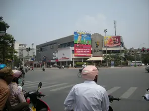 Bối cảnh là một ngã tư đường phố đông đúc ở Hà Nội vào ban ngày. Nhiều người đang điều khiển xe máy và một số đi xe đạp, dừng lại chờ đèn tín hiệu giao thông. Hầu hết mọi người đều đội mũ bảo hiểm; một người ở phía trước đội mũ bảo hiểm màu hồng có chữ "Scramble".。Ở phía đối diện của ngã tư là một tòa nhà thương mại lớn. Trên tòa nhà có nhiều biển quảng cáo và biển hiệu:。- Biển quảng cáo lớn màu đỏ của "PICO PLAZA", được mô tả là "HỆ THỐNG SIÊU THỊ ĐIỆN MÁY HÀNG ĐẦU VIỆT NAM".。- Phía trên là biển quảng cáo cho nước giải khát C2 với dòng chữ "mát lạnh & mạnh khiết".。- Bên phải là một biển quảng cáo khác có hình một nhóm người mặc áo đỏ.。- Tên của tòa nhà dường như là "HANOI SUPERBOWL", có thể nhìn thấy bên dưới các biển quảng cáo.。- Ở tầng trệt, có một cửa hàng với biển hiệu của nhà mạng "S-fone".。- Ở phía bên trái của bức ảnh, có một biển hiệu bị che một phần ghi "VIỆN GIA SƯ".。Cảnh vật xung quanh bao gồm các cột đèn giao thông, vạch kẻ đường dành cho người đi bộ, cây cối ven đường, và một cột tháp viễn thông trên nóc tòa nhà. Một chiếc xe tải nhỏ màu trắng cũng xuất hiện ở phía xa.。（日本語訳）。背景は、日中のハノイにある混雑した交差点です。多くの人々がバイクを運転し、何人かは自転車に乗って、交通信号を待って停止しています。ほとんどの人はヘルメットを着用しています。前方の一人は「Scramble」という文字が入ったピンク色のヘルメットをかぶっています。交差点の向かい側には大きな商業ビルがあります。そのビルには多くの広告看板や標識があります：。- 「PICO PLAZA」の大きな赤い広告看板で、「HỆ THỐNG SIÊU THỊ ĐIỆN MÁY HÀNG ĐẦU VIỆT NAM」（ベトナムトップの家電スーパーマーケットシステム）と説明されています。- その上には、清涼飲料水C2の広告看板があり、「mát lạnh & mạnh khiết」という文言が書かれています。- 右側には、赤い服を着た人々のグループが描かれた別の広告看板があります。- ビルの名前は「HANOI SUPERBOWL」のようで、広告看板の下に見ることができます。- 1階には、通信事業者「S-fone」の看板がある店舗があります。- 写真の左側には、「VIỆN GIA SƯ」と書かれた、部分的に隠れている看板があります。周囲の光景には、信号機、横断歩道、街路樹、そしてビルの屋上にある通信塔が含まれます。遠くには白い小型トラックも見えます。