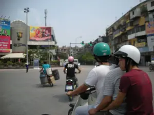 Bức ảnh chụp cảnh giao thông tại một ngã tư đường phố đông đúc ở một thành phố, có thể là Hà Nội. Có nhiều xe máy đang di chuyển trên đường.。Về chữ viết, có thể nhận dạng một số nội dung trên các biển quảng cáo và vật thể:。- Tòa nhà bên trái có biển hiệu lớn màu đỏ và hồng với các dòng chữ "PLAZA", "ĐIỆN MÁY", "VIỆT NAM".。- Bên cạnh đó là một biển quảng cáo màu xanh lá cây có chữ "...lành & thanh khiết", phần còn lại không rõ.。- Một người đi xe máy mặc áo phông đen có số "501" ở sau lưng.。- Các biển hiệu và chữ viết khác bị mờ, không thể đọc rõ nội dung.。Về tình cảnh và hoạt động:。- **Loại địa điểm:** Ngã tư đường phố đô thị với nhiều tòa nhà cao tầng, bao gồm cả các tòa nhà hiện đại và các khu nhà tập thể cũ đặc trưng. Có cột đèn giao thông đang bật tín hiệu màu xanh.。- **Hoạt động cụ thể:** Cảnh tượng giao thông đường phố điển hình. Ở tiền cảnh, ba thanh niên đang đi chung một chiếc xe máy. Người lái đội mũ bảo hiểm màu xanh lá cây, người ngồi giữa mặc áo phông kẻ sọc, và người ngồi sau cùng mặc áo phông màu đỏ và đội mũ bảo hiểm màu trắng. Một người khác đang lái xe máy chở hàng cồng kềnh, với nhiều thùng lớn được buộc ở hai bên. Ở phía xa, có một người đi bộ đội nón lá.。- **Vật thể trong ảnh:** Phương tiện giao thông chủ yếu là xe máy (xe máy). Hầu hết người tham gia giao thông đều đội mũ bảo hiểm (mũ bảo hiểm). Ngoài ra còn có các tòa nhà, biển quảng cáo lớn, cột đèn và đèn tín hiệu giao thông.。（日本語訳）。ある都市、おそらくハノイの、混雑した街の交差点での交通風景を撮影した写真です。道路上では多くのバイクが移動しています。文字については、広告看板や物体の上にあるいくつかの内容を認識できます。- 左側の建物には、「PLAZA」、「ĐIỆN MÁY」（家電）、「VIỆT NAM」（ベトナム）という文字が書かれた大きな赤とピンクの看板があります。- その隣には、「...lành & thanh khiết」（...健康的＆純粋）という文字がある緑色の広告看板がありますが、残りの部分は不明瞭です。- バイクに乗っている一人は、背中に「501」という数字が入った黒いTシャツを着ています。状況と活動について：。- **場所の種類：** 多くの高層ビルがある都市の街の交差点で、近代的なビルと特徴的な古い集合住宅の両方が含まれています。信号機が青信号を灯しています。- **具体的な活動：** 典型的な街の交通風景。前景では、3人の若者が1台のバイクに相乗りしています。運転手は緑色のヘルメットをかぶり、真ん中に座っている人はストライプのTシャツを着ており、一番後ろに座っている人は赤いTシャツを着て白いヘルメットをかぶっています。別の人は、両側に大きな箱をいくつも縛り付けたかさばる荷物を運んでバイクを運転しています。遠くには、ノンラー（菅笠）をかぶった歩行者が一人います。- **写真の中の物体：** 主な交通手段はバイク（xe máy）です。交通参加者のほとんどはヘルメット（mũ bảo hiểm）を着用しています。その他に、建物、大きな広告看板、電柱、信号機があります。