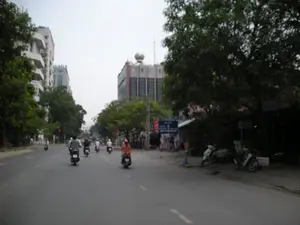 Quang cảnh một con đường đô thị ở Hà Nội với nhiều xe máy đang lưu thông. Một số người đang dừng xe chờ đèn đỏ. Hai bên đường có nhiều cây xanh lớn. Phía bên trái là các tòa nhà cao tầng hiện đại. Phía bên phải là vỉa hè với một số quán nhỏ ven đường và vài chiếc xe máy đang đậu. Có một biển báo giao thông màu xanh lam chỉ hướng nhưng chữ viết bị mờ không đọc được. Ở phía xa, có thể nhìn thấy các tòa nhà cao tầng khác, một trong số đó có kiến trúc khá đặc biệt ở phần mái.。（日本語訳）。ハノイのある都市部の道路の光景で、多くのバイクが通行しています。何人かの人々が赤信号を待って停車しています。道路の両側には多くの大きな緑の木があります。左側には近代的な高層ビルがあります。右側は歩道で、いくつかの小さな道端の店と数台のバイクが駐車しています。方向を示す青い交通標識がありますが、文字がぼやけていて読めません。遠くには他の高層ビルが見え、そのうちの一つは屋根の部分にかなり特徴的な建築様式を持っています。