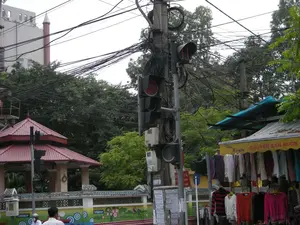 Bản ghi văn bản: Trên biển hiệu màu vàng của một cửa hàng quần áo có ghi: "Thu Huyền" và "CHUYÊN BÁN BUÔN". Trên cột điện có dán nhiều tờ rơi, áp phích nhưng chữ quá nhỏ và mờ không thể đọc rõ nội dung. Có một biển báo nhỏ màu đỏ và trắng trên cột điện, có thể đọc được chữ "CA" và một phần số điện thoại. Trên bức tường trang trí bên trái có những chữ viết cách điệu, nhưng không rõ nghĩa.。Mô tả cảnh quan và hoạt động: Bức ảnh chụp một góc phố đô thị ở Việt Nam, có thể là Hà Nội. Điểm nổi bật nhất là một cột điện bê tông ở trung tâm với một mớ dây điện và cáp viễn thông chằng chịt, rối tung. Trên cột có gắn nhiều cụm đèn tín hiệu giao thông, trong đó có một đèn đang báo màu đỏ, cho thấy hoạt động điều tiết giao thông. Bên phải là một cửa hàng quần áo vỉa hè, có mái che bằng bạt, đang treo bán nhiều loại áo sơ mi và áo len. Bên trái là một công trình kiến trúc nhỏ có mái ngói đỏ theo kiểu truyền thống, có thể là một vọng lâu hoặc cổng chào. Bên dưới là một bức tường được trang trí bằng các họa tiết và tranh vẽ nhiều màu sắc. Phía sau là cây cối xanh tốt và một phần của một tòa nhà cao tầng. Ở góc dưới bên trái có hai người đang ở trên đường, một người đội mũ bảo hiểm.。（日本語訳）。テキスト記録：ある衣料品店の黄色い看板には、「Thu Huyền」と「CHUYÊN BÁN BUÔN（卸売専門）」と書かれています。電柱には赤と白の小さな看板があり、「CA」という文字と電話番号の一部が読み取れます。左側の装飾壁にはデザイン化された文字がありますが、意味は不明です。風景と活動の説明：写真はベトナムの都市の街角を撮影したもので、ハノイかもしれません。最も目立つのは、中央にあるコンクリートの電柱で、無数の電線や通信ケーブルが複雑に絡み合っています。柱には複数の交通信号機が設置されており、そのうちの一つは赤色を点灯しており、交通規制が行われていることを示しています。右側には歩道にある衣料品店があり、防水シートの屋根があり、多くの種類のシャツやセーターを吊るして販売しています。左側には伝統的な様式の赤い瓦屋根を持つ小さな建築物があり、物見やぐらか歓迎の門かもしれません。その下には、色とりどりの模様や絵で装飾された壁があります。後方には青々とした木々と高層ビルの一部が見えます。左下の角には道路上に二人の人物がおり、一人はヘルメットをかぶっています。
