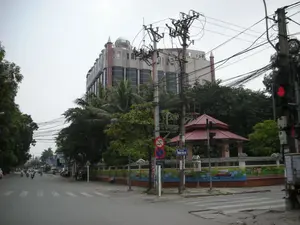 Bức ảnh chụp một góc phố tại một ngã tư ở khu vực đô thị. Có một biển báo tên đường màu xanh ghi chữ 'PHỐ VĂN TRÌ'. Bên dưới là biển báo giao thông hình tròn cấm dừng và cấm đỗ xe. Ở phía sau, nổi bật là một tòa nhà lớn, cao tầng với kiến trúc độc đáo, có phần mái vòm trên đỉnh. Trên mái vòm có chữ nhưng không thể đọc rõ. Ở góc ngã tư là một công trình kiến trúc nhỏ kiểu nhà vọng lâu với mái ngói đỏ, được bao quanh bởi một bức tường được trang trí bằng những bức vẽ sặc sỡ. Trên đường có nhiều người đang đi xe máy, thể hiện hoạt động giao thông thường ngày. Hệ thống cột điện với mạng lưới dây điện và dây cáp chằng chịt là một đặc điểm nổi bật của khung cảnh. Cây xanh, bao gồm cả cây cọ, được trồng dọc hai bên đường. Một cột đèn tín hiệu giao thông đang hiển thị màu đỏ.。（日本語訳）。写真は都市部のある交差点の街角を撮影したものです。「PHỐ VĂN TRÌ」という文字が書かれた青い通りの名前の標識があります。その下には、駐停車禁止の円形の交通標識があります。後方には、頂上にドーム屋根を持つユニークな建築様式の大きな高層ビルが際立っています。ドーム屋根には文字がありますが、はっきりと読むことはできません。交差点の角には、赤い瓦屋根の望楼風の小さな建築物があり、色鮮やかな絵で装飾された壁に囲まれています。道路上では多くの人がバイクに乗っており、日常の交通活動を示しています。複雑に絡み合った電線とケーブル網を持つ電柱システムは、この風景の際立った特徴です。ヤシの木を含む緑の木々が、道路の両脇に植えられています。交通信号機が赤色を表示しています。