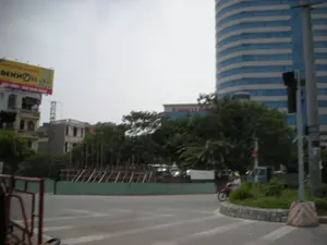 Trên tòa nhà bên trái có một biển quảng cáo lớn màu vàng. Dòng chữ có thể đọc được một phần là 'DEKHN D25' và 'SANG TRỌNG - ĐẲNG CẤP SỐNG'. Phía sau hàng cây, trên một tòa nhà khác, có một tấm biển với dòng chữ dường như là '... CÔNG TY ...', nhưng không thể đọc rõ toàn bộ.。Đây là khung cảnh tại một ngã tư hoặc vòng xuyến ở một khu đô thị tại Hà Nội. Phía bên phải là một tòa nhà cao tầng hiện đại, có mặt tiền bằng kính màu xanh và hình dáng cong. Phía bên trái là các tòa nhà phố cũ hơn. Ở giữa là một khu đất trống được rào lại bằng hàng rào tôn màu xanh, bên trong có các cọc gỗ, cho thấy đây là một công trường đang thi công. Một người đang lái xe máy màu đỏ đi trên đường. Ngoài ra còn có các vật thể khác như cột đèn giao thông, cây xanh và một khối bê tông sơn sọc trắng đỏ ở lề đường.。（日本語訳）。左側の建物には、大きな黄色い広告看板があります。部分的に読み取れる文字は「DEKHN D25」と「SANG TRỌNG - ĐẲNG CẤP SỐNG」です。これはハノイのある都市部の交差点またはロータリーでの光景です。右側には、青いガラスのファサードを持ち、湾曲した形状の近代的な高層ビルがあります。左側には、より古いタウンハウスがあります。中央には青いトタンのフェンスで囲まれた空き地があり、内部には木の杭があって、ここが工事中の現場であることを示しています。一人の人が赤いバイクを運転して道路を走っています。その他にも、信号機、緑の木々、そして路肩にある赤と白の縞模様に塗られたコンクリートブロックなどの物体があります。
