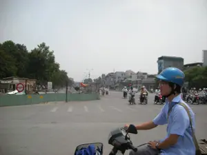 Bối cảnh là một con đường nhựa tại một thành phố, có thể là Hà Nội, với nhiều người đang tham gia giao thông bằng xe máy. Ở phía trước, một người đàn ông trẻ tuổi mặc áo sơ mi xanh, đeo kính và đội mũ bảo hiểm màu xanh dương đang điều khiển một chiếc xe máy. Phía sau anh là dòng xe máy đông đúc, một hình ảnh đặc trưng của giao thông đô thị Việt Nam. Bên trái đường là một khu vực công trường đang được thi công, được quây lại bằng hàng rào tôn màu xanh lá cây. Trên hàng rào có một số biển báo. Một biển báo hình tròn màu đỏ với vạch trắng ngang ở giữa (biển báo cấm đi vào). Một biển báo khác màu trắng chữ đỏ có vẻ là biển cảnh báo công trường nhưng chữ đã bị mờ, khó đọc. Phía sau hàng rào là cây cối và một căn nhà tạm. Xa hơn trong hậu cảnh là những tòa nhà phố san sát nhau.。（日本語訳）。背景は、ある都市、おそらくハノイのアスファルト道路で、多くの人々がバイクで交通に参加しています。前方には、青いシャツを着て、眼鏡をかけ、青いヘルメットをかぶった若い男性がバイクを運転しています。彼の後ろには混雑したバイクの流れがあり、ベトナムの都市交通の典型的な光景です。道路の左側には工事中の現場があり、緑色のトタンのフェンスで囲われています。フェンスにはいくつかの標識があります。一つは、中央に白い横線が入った赤色の円形標識（進入禁止標識）です。もう一つの標識は白地に赤文字で、工事現場の警告標識のようですが、文字がかすれていて読みにくいです。フェンスの後ろには木々と仮設の家があります。さらに遠くの背景には、商店が密集しています。