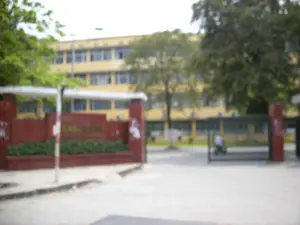 Bức ảnh chụp lối vào của một tòa nhà lớn, có thể là một trường học, trường đại học hoặc một cơ quan hành chính. Ở phía sau là một tòa nhà nhiều tầng màu vàng với nhiều cửa sổ. Phía trước là cổng vào được xây bằng gạch màu nâu đỏ, trên đó có gắn chữ màu vàng nhưng không thể đọc được do ảnh bị mờ. Bên cạnh là một cánh cổng sắt màu đen. Có một người, dường như là nhân viên bảo vệ, đang ngồi ở khu vực cổng. Xung quanh có nhiều cây xanh.。（日本語訳）。大きな建物の入り口を写した写真で、学校、大学、または行政機関である可能性があります。後方には、多くの窓がある黄色い多層階の建物があります。手前には赤褐色のレンガで造られた門があり、その上には黄色の文字が取り付けられていますが、写真がぼやけているため読むことはできません。その隣には黒い鉄の門があります。警備員と思われる人物が一人、門のエリアに座っています。周囲には多くの緑の木々があります。