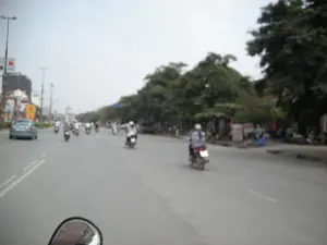 Bức ảnh mờ chụp lại cảnh giao thông trên một con đường rộng ở Hà Nội. Có rất nhiều người đang điều khiển xe máy di chuyển trên đường. Ở phía bên trái, có một chiếc ô tô đang chạy. Dọc theo lề đường bên phải là hàng cây xanh lớn. Ở phía xa có thể nhìn thấy các tòa nhà, cột đèn đường và các biển báo, biển quảng cáo nhưng tất cả đều bị mờ và không thể đọc được chữ. Góc dưới của bức ảnh cho thấy một phần gương chiếu hậu của phương tiện mà người chụp đang sử dụng, có thể là một chiếc xe máy.。（日本語訳）。ハノイの広い道路上の交通風景を撮影したぼやけた写真です。多くの人々がバイクを運転して道路を移動しています。左側には、一台の車が走っています。右側の路肩に沿って、大きな緑の木々の列があります。遠くには建物、街灯、標識や広告看板が見えますが、すべてぼやけており、文字を読むことはできません。写真の下隅には、撮影者が使用している乗り物のバックミラーの一部が写っており、それはバイクである可能性があります。