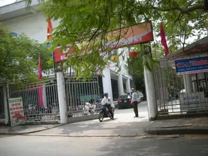 Bức ảnh chụp quang cảnh tại cổng vào của một tòa nhà hoặc một khu phức hợp trên một con phố ở Hà Nội. Cảnh vật diễn ra dưới bóng cây xanh rậm rạp. Một người đàn ông đội mũ bảo hiểm đang lái xe máy đi qua cổng sắt đang mở. Một người đàn ông khác đội mũ rộng vành đứng ở trong sân, có thể là nhân viên bảo vệ. Bên trong sân, có một chiếc ô tô màu đen và nhiều xe máy khác đang được gửi. Tòa nhà phía sau có kiến trúc hiện đại với các cột trụ màu trắng. Có nhiều biển hiệu và cờ được treo lên.。Nội dung các văn bản có thể đọc được:。1. Biển hiệu lớn màu đỏ phía trên cổng: Chữ bị cành cây che khuất nhiều, chỉ đọc được một phần "... LỚP THU IV".。2. Biển hiệu màu xanh bên phải cổng: "Anh ngữ London", "www.london.edu.vn", "ANH VĂN - TIN HỌC CHẤT LƯỢNG CAO". Đây là biển hiệu của một trung tâm tiếng Anh.。3. Biển hiệu màu xanh lá cây bên trái, đặt trên vỉa hè: "NHÀ HÀNG DUYÊN QUÊ", "CƠM VIỆT NAM", "HẢI SẢN TƯƠI SỐNG", "CƠM CHÁY BẮC BỘ". Đây là biển quảng cáo cho một nhà hàng.。4. Biển báo nhỏ bên trong cổng: Có chữ "NƠI GỬI XE...", cho biết đây là khu vực giữ xe.。Ngoài ra, có các lá cờ đỏ và đèn lồng màu đỏ, vàng được treo trang trí.。（日本語訳）。写真は、ハノイのある通りにある建物または複合施設の入り口の光景を撮影したものです。光景は、青々と茂った木々の木陰で繰り広げられています。ヘルメットをかぶった一人の男性が、開いている鉄の門を通り抜けてバイクを運転しています。もう一人の男性が、つばの広い帽子をかぶって敷地内に立っており、おそらく警備員でしょう。敷地内には、一台の黒い車と他の多くのバイクが駐車されています。後ろの建物は現代的な建築様式で、白い柱があります。多くの看板と旗が掲げられています。読み取れるテキストの内容：。1. 門の上にある大きな赤い看板：文字は木の枝で多くが隠されており、「... LỚP THU IV」という一部分しか読めません。2. 門の右側にある青い看板：「Anh ngữ London」、「www.london.edu.vn」、「ANH VĂN - TIN HỌC CHẤT LƯỢNG CAO」。これは英語センターの看板です。3. 左側の歩道に置かれた緑色の看板：「NHÀ HÀNG DUYÊN QUÊ」、「CƠM VIỆT NAM」、「HẢI SẢN TƯƠI SỐNG」、「CƠM CHÁY BẮC BỘ」。これはレストランの広告看板です。4. 門の内側にある小さな標識：「NƠI GỬI XE...」という文字があり、ここが駐輪場であることを示しています。その他に、赤い旗と赤と黄色の提灯が飾りに吊るされています。