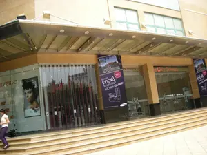 Bức ảnh chụp lối vào của một tòa nhà hiện đại, có vẻ là một trung tâm thương mại hoặc tòa nhà văn phòng ở Hà Nội. Phía trên cửa ra vào chính bằng kính có dòng chữ 'Hanoi City Towers'. Lối vào có một cầu thang bộ rộng và một mái hiên lớn bằng kính và kim loại. Về phía bên trái là mặt tiền của một cửa hàng với cửa kính lớn, trưng bày áp phích quảng cáo của thương hiệu mỹ phẩm 'SHISEIDO'. Ở khu vực trung tâm có treo một biểu ngữ quảng cáo lớn màu tím cho điện thoại di động 'Samsung innov8'. Nội dung trên biểu ngữ ghi: 'Samsung innov8', 'TỰ HÀO CHỤP BỨC HÌNH LỚN NHẤT'. Nhìn qua lớp cửa kính, có thể thấy nhiều lá cờ đỏ sao vàng của Việt Nam được trưng bày bên trong. Một người phụ nữ mặc áo trắng và quần tím đang đi lên cầu thang ở phía bên trái. Một chiếc xe máy đang đậu gần cửa hàng Shiseido.。（日本語訳）。写真は、ハノイにある近代的な建物の入り口を撮影したもので、ショッピングセンターかオフィスビルのようです。ガラス張りのメインエントランスの上には「Hanoi City Towers」という文字があります。入り口には幅の広い階段と、ガラスと金属でできた大きな庇があります。左側には、大きなガラス扉のある店舗のファサードがあり、化粧品ブランド「SHISEIDO」の広告ポスターが飾られています。中央エリアには、携帯電話「Samsung innov8」の紫色の大きな広告バナーが掲げられています。バナーには「Samsung innov8」、「TỰ HÀO CHỤP BỨC HÌNH LỚN NHẤT（最大の写真を撮る誇り）」と書かれています。ガラス扉越しに、中には多くのベトナムの金星紅旗が飾られているのが見えます。白いシャツと紫のズボンを履いた女性が、左側の階段を上っています。資生堂の店の近くにバイクが一台停まっています。