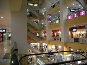 Đây là hình ảnh bên trong một trung tâm thương mại lớn và hiện đại có nhiều tầng. Khung cảnh được chụp từ một tầng cao, nhìn ra khu vực sảnh chính (giếng trời), cho thấy nhiều tầng được kết nối với nhau bằng hệ thống thang cuốn chéo. Kiến trúc nổi bật với các cột trụ lớn màu trắng, lan can và tường màu vàng, cùng sàn nhà lát gạch sáng bóng.。Các hoạt động diễn ra chủ yếu là mua sắm và đi dạo. Có thể thấy nhiều người đang di chuyển trên các tầng, một số đi trên thang cuốn, một số đi dọc hành lang và một số khác đang ở trong các cửa hàng.。Nhiều biển hiệu và chữ viết có thể được nhận dạng:。- Tên siêu thị "Citimart" trên một biển hiệu lớn màu đỏ ở tầng trên.。- Logo của thương hiệu "LG" xuất hiện nhiều lần. Một biển quảng cáo cụ thể ghi "scarlet LG LCD TV 2008".。- Tên các thương hiệu cao cấp khác có thể nhìn thấy trên các cửa hàng, bao gồm "L'OCCITANE", "RADO", và một phần của "ARMANI" và "VERSACE".。- Nhiều màn hình TV đang hoạt động, có thể đang chiếu quảng cáo.。- Có nhiều biển hiệu khác nhưng không thể đọc được do mờ hoặc ở khoảng cách xa.。（日本語訳）。これは、多くの階層を持つ、大きくて近代的なショッピングセンターの内部の画像です。景色は高い階から撮影されており、メインホール（吹き抜け）を見下ろしています。多くの階が斜めに交差するエスカレーターシステムで接続されているのがわかります。建築は、大きな白い柱、黄色い手すりと壁、そして光沢のあるタイル張りの床が特徴です。主に行われている活動は、買い物と散策です。多くの人々が各階を移動しているのが見えます。エスカレーターに乗っている人、廊下を歩いている人、そして店の中にいる人もいます。多くの看板や文字が認識できます：。- 上の階にある大きな赤い看板に書かれたスーパーマーケット名「Citimart」。- 「LG」ブランドのロゴが何度も表示されています。特定の広告看板には「scarlet LG LCD TV 2008」と書かれています。- 店舗には、「L'OCCITANE」、「RADO」、そして「ARMANI」と「VERSACE」の一部など、他の高級ブランド名が見えます。- 多くのテレビ画面が作動しており、おそらく広告を放映しています。- 他にも多くの看板がありますが、ぼやけていたり、遠距離にあるため読み取れません。