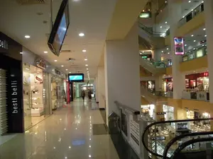 Bức ảnh chụp bên trong một trung tâm thương mại hiện đại, nhiều tầng. Không gian là một hành lang rộng rãi với sàn gạch sáng bóng. Dọc theo hành lang bên trái là các cửa hàng có mặt tiền bằng kính. Một cửa hàng có tên thương hiệu 'besti belli' được viết dọc bằng chữ trắng trên một cột màu đen. Các cửa hàng khác cũng có biển hiệu nhưng chữ viết không rõ ràng. Ở phía xa, có một vài người đang đi bộ. Có hai màn hình TV được treo trên trần nhà. Nhìn sang bên phải là một không gian mở lớn với nhiều tầng của trung tâm thương mại, được kết nối với nhau bằng thang cuốn. Có thể thấy các biển quảng cáo và logo thương hiệu, bao gồm cả logo 'LG' trên một tầng cao hơn. Toàn bộ không gian được chiếu sáng rực rỡ bằng hệ thống đèn âm trần.。（日本語訳）。現代的な多層階のショッピングモール内で撮影された写真です。空間は、光沢のあるタイル張りの床が特徴の広々とした廊下です。左側の廊下に沿って、ガラス張りの店舗が並んでいます。ある店舗には、黒い柱に白文字で縦に「besti belli」というブランド名が書かれています。他の店舗にも看板がありますが、文字ははっきりと見えません。遠くには、数人の人が歩いています。天井から2台のテレビモニターが吊り下げられています。右側を見ると、エスカレーターで結ばれたショッピングモールの複数階にわたる大きな吹き抜け空間があります。上の階にある「LG」のロゴをはじめ、広告看板やブランドロゴが見えます。空間全体が、埋め込み式の天井照明システムによって明るく照らされています。