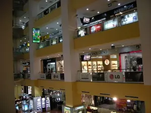 Bức ảnh chụp không gian bên trong một trung tâm thương mại hiện đại và nhiều tầng ở Hà Nội. Khung cảnh được chụp từ một tầng cao, nhìn xuống và ngang qua khu vực giếng trời trung tâm, cho thấy kiến trúc với nhiều tầng lầu và các cửa hàng san sát.。Các chữ viết và biển hiệu có thể nhận dạng bao gồm:。- Tên các cửa hàng và thương hiệu: AKEMI UCHI, WORLD KITCHEN, ROMANSON, ROAMER, CANDINO, VICENZO ZENO, besti belli.。- Thương hiệu điện tử: LG, logo xuất hiện trên các biểu ngữ quảng cáo dọc theo lan can.。- Quảng cáo: Một màn hình kỹ thuật số dạng dọc đang treo ở khu vực giếng trời. Các tấm biển quảng cáo lớn cho các thương hiệu đồng hồ và sản phẩm của LG (EDGE).。- Chữ tiếng Việt: Một biểu ngữ ở tầng dưới có ghi 'DỰ THƯỞNG' và 'GIẢI THƯỞNG LỚN'.。Hoạt động và cảnh quan:。- Nơi chốn: Đây là một trung tâm thương mại hoặc khu mua sắm phức hợp.。- Hoạt động: Một vài người đang đi lại trên các hành lang và bên trong các cửa hàng, có thể đang mua sắm hoặc tham quan.。- Vật thể: Nhiều cửa hàng với mặt tiền bằng kính, các kệ trưng bày sản phẩm (giày dép trong cửa hàng VICENZO ZENO), ma-nơ-canh trong cửa hàng thời trang, hệ thống đèn chiếu sáng trên trần, lan can bằng kính và kim loại, và thang cuốn có thể thấy ở phía xa. Toàn bộ không gian được chiếu sáng rực rỡ.。（日本語訳）。ハノイにある、モダンで多層階のショッピングモールの内部空間を撮影した写真です。この光景は高層階から撮影されており、中央の吹き抜けエリアを見下ろし、横切るように見ています。多くのフロアと密集した店舗がある建築様式が見て取れます。識別可能な文字や看板には、以下のものが含まれます。- 店舗名およびブランド名：AKEMI UCHI, WORLD KITCHEN, ROMANSON, ROAMER, CANDINO, VICENZO ZENO, besti belli。- 電子機器ブランド：LG、ロゴが手すりに沿った広告バナーに表示されています。- 広告：吹き抜けエリアに縦型のデジタルスクリーンが吊り下げられています。時計ブランドやLG製品（EDGE）の大きな広告看板があります。- ベトナム語の文字：下の階のバナーに「DỰ THƯỞNG」（懸賞）と「GIẢI THƯỞNG LỚN」（大賞）と書かれています。活動と景観：。- 場所：ここはショッピングモールまたは複合商業施設です。- 活動：数人が廊下や店舗内を歩き回っており、買い物をしたり見て回ったりしているようです。空間全体が明るく照らされています。