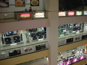 Đây là quang cảnh bên trong một trung tâm thương mại hoặc cửa hàng bách hóa nhiều tầng, được chụp từ trên cao nhìn xuống. Không gian được chia thành nhiều tầng với lan can bằng kính. Các tầng giữa chủ yếu là khu vực kinh doanh đồ điện tử, trưng bày rất nhiều tivi màn hình phẳng. Có thể nhận thấy rõ các biển hiệu của những thương hiệu lớn như "SHARP" (với dòng sản phẩm "AQUOS"), "LG", "JVC", và "Panasonic". Một biển quảng cáo đáng chú ý là "scarlet LG LCD TV 2008". Ở tầng trên cùng, có một quầy bán đồ ăn mang tên "DELI MANJOO" với các biển hiệu đèn và hình ảnh các món ăn. Ở một trong các tầng dưới, có một gian hàng bày bán nhiều loại mũ, nơi một vài người đang xem hàng. Khung cảnh chung cho thấy hoạt động mua sắm và tham quan trong một không gian thương mại hiện đại.。（日本語訳）。これは多層階のショッピングセンターまたはデパートの内部の光景で、上から見下ろして撮影されたものです。空間はガラスの手すりがある多くの階に分かれています。中間の階は主に電化製品の販売エリアで、非常に多くの薄型テレビが展示されています。「SHARP」（製品ライン「AQUOS」）、「LG」、「JVC」、「Panasonic」といった大手ブランドの看板がはっきりと見て取れます。注目すべき広告看板の一つは「scarlet LG LCD TV 2008」です。最上階には、「DELI MANJOO」という名前の食べ物の売店があり、電飾看板と料理の写真が掲げられています。下の階の一つには、多くの種類の帽子を陳列販売しているブースがあり、数人が商品を見ています。全体的な光景は、現代的な商業空間での買い物や見て回る活動を示しています。