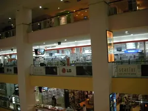 Đây là hình ảnh chụp bên trong một trung tâm thương mại hoặc siêu thị điện máy lớn có nhiều tầng. Không gian được chiếu sáng tốt với nhiều đèn trần. Bức ảnh cho thấy các tầng lầu khác nhau với lan can bằng kính, nhìn ra một không gian chung ở giữa. Các cửa hàng chủ yếu kinh doanh các mặt hàng điện tử, với nhiều TV màn hình phẳng được trưng bày. Một số người, có thể là khách hàng và nhân viên, đang có mặt tại các tầng. Có thể đọc được một số văn bản từ các biển hiệu và băng rôn quảng cáo: tên cửa hàng 'BEST CARINGS', quảng cáo cho TV 'scarlet LG LCD TV 2008' cùng với logo 'LG'. Ngoài ra, có thể nhận ra các biển hiệu của các thương hiệu khác như 'Panasonic' và 'TOSHIBA' bên trong các gian hàng.。（日本語訳）。これは、複数階建ての大きなショッピングセンターまたは家電量販店の内部で撮影された写真です。空間は多くの天井照明で明るく照らされています。写真には、ガラスの手すりが付いたさまざまなフロアが写っており、中央の共有スペースを見下ろしています。店舗は主に電子製品を取り扱っており、多くの薄型テレビが展示されています。各階には、客や従業員と思われる数人の人々がいます。看板や広告バナーからいくつかの文字を読み取ることができます。店名は「BEST CARINGS」、テレビの広告は「scarlet LG LCD TV 2008」で、「LG」のロゴが付いています。さらに、各ブース内には「Panasonic」や「TOSHIBA」といった他のブランドの看板も認識できます。