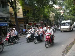 Bức ảnh ghi lại cảnh giao thông đông đúc trên một con phố ở Hà Nội. Dọc hai bên đường là những tòa nhà ống cao tầng đặc trưng, với tầng trệt được sử dụng làm cửa hàng. Hoạt động chính là mọi người đang tham gia giao thông, chủ yếu bằng xe máy. Rất nhiều người, cả nam và nữ, đang điều khiển xe máy và tất cả đều đội mũ bảo hiểm. Có thể thấy một gia đình gồm cha mẹ và một đứa trẻ nhỏ cùng đi trên một chiếc xe. Ngoài xe máy, còn có một chiếc xe khách loại nhỏ (xe van) màu trắng ngà đang lưu thông. Ven đường có nhiều cây xanh lớn. Một số biển hiệu cửa hàng có thể được nhìn thấy, bao gồm một biển hiệu màu xanh với dòng chữ "LỮU ĐẶC BIỆT", và các phần của các biển hiệu khác như "...CAMERA" và "...CABENA". Các biển hiệu còn lại bị che khuất hoặc không thể đọc rõ.。（日本語訳）。写真はハノイのある通りでの混雑した交通風景を捉えています。道の両脇には、特徴的な高層のチューブハウスが並び、1階は店舗として使われています。主な活動は、人々が主にバイクで交通に参加していることです。多くの人々、男性も女性も、バイクを運転しており、全員がヘルメットを着用しています。両親と小さな子供からなる家族が1台のバイクに一緒に乗っているのが見えます。バイクの他に、アイボリー色の小型バス（バン）も走行しています。道端には大きな木が多くあります。いくつかの店の看板が見え、その中には「LỮU ĐẶC BIỆT」という文字が書かれた青い看板や、「...CAMERA」や「...CABENA」のような他の看板の一部が含まれています。残りの看板は隠れているか、はっきりと読み取れません。