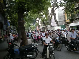 Bối cảnh là một con phố đô thị đông đúc ở Hà Nội, với dòng xe cộ tấp nập. Hoạt động chính là giao thông đường phố, với rất nhiều người đang điều khiển xe máy, tạo nên cảnh tượng có vẻ như là ùn tắc hoặc di chuyển chậm. Hầu hết mọi người đi xe máy đều đội mũ bảo hiểm, một số người còn đeo khẩu trang.。Hai bên đường là những hàng cây xanh lớn và các tòa nhà nhiều tầng san sát nhau, tầng trệt có vẻ là các cửa hàng. Phía bên trái, có thể thấy những tấm bạt màu xanh của các cửa hàng nhưng chữ viết trên đó không thể đọc rõ. Hệ thống dây điện chằng chịt treo phía trên bên trái con đường, một cảnh tượng thường thấy ở các khu đô thị Việt Nam.。Ở phía bên phải của bức ảnh, có một chiếc xe buýt nhỏ màu trắng với dòng chữ có thể đọc được là "HUY HÙNG 131 BA". Nhiều loại xe máy khác nhau có thể được nhìn thấy trên đường. Một số người đang đứng trên vỉa hè, trong khi những người khác ngồi trên ghế nhựa màu xanh.。（日本語訳）。背景はハノイの混雑した都市の通りで、交通量が非常に多いです。バイクに乗っているほとんどの人はヘルメットを着用しており、マスクを着用している人もいます。道路の両側には大きな緑の並木と密集した多層階の建物があり、1階は店舗のようです。左側には、店舗の青いタープが見えますが、そこに書かれている文字ははっきりと読めません。絡み合った電線が道路の左上にぶら下がっており、これはベトナムの都市部でよく見られる光景です。写真の右側には、白い小型バスがあり、「HUY HÙNG 131 BA」と読める文字が書かれています。様々な種類のバイクが道路上に見られます。歩道に立っている人もいれば、青いプラスチックの椅子に座っている人もいます。