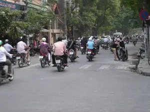 Về chữ viết: Có một tấm biển hiệu của cửa hàng ở bên trái với dòng chữ màu trắng trên nền xanh: 'SỬA MÁY MAY QUẦN ÁO'. Ở bên phải đường có một biển báo giao thông hình tròn, viền đỏ, nền xanh với một gạch chéo đỏ, là biển báo cấm dừng xe.。Về khung cảnh và hoạt động: Bức ảnh chụp một con đường phố tại Hà Nội, với mật độ giao thông đông đúc, chủ yếu là xe máy (xe gắn máy). Rất nhiều người đang điều khiển xe máy trên đường, hầu hết đều đội mũ bảo hiểm. Khung cảnh cho thấy một dòng người và xe đang di chuyển. Ở phía bên phải, một cặp đôi đang đứng trên vỉa hè bên cạnh chiếc xe máy của họ. Hai bên đường là các tòa nhà nhiều tầng và cây xanh. Trên mặt đường có vạch kẻ trắng dành cho người đi bộ.。（日本語訳）。文字について：左側には店の看板が一つあり、青い背景に白い文字で「SỬA MÁY MAY QUẦN ÁO」と書かれています。道路の右側には、円形で赤枠、青地に赤い斜線が入った交通標識があり、これは停車禁止の標識です。風景と活動について：写真はハノイのある通りを撮影したもので、交通量が多く、主にバイク（原動機付自転車）です。たくさんの人が道路でバイクを運転しており、そのほとんどがヘルメットを着用しています。その光景は、移動している人々と車両の流れを示しています。右側では、一組のカップルが歩道上で自分たちのバイクの隣に立っています。道路の両側には、複数階建ての建物と緑の木々があります。路面には歩行者用の白い線が引かれています。