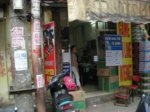 Bối cảnh là một con phố ở Hà Nội, chụp phía trước một quán Internet và game. Đây là một cửa hàng nhỏ, mở thẳng ra vỉa hè, có mái hiên bạt đã cũ. Bên trong quán, có thể thấy nhiều màn hình máy tính và một vài người đang ngồi, có lẽ là khách hàng đang chơi game hoặc sử dụng Internet. Một người phụ nữ đang đứng ở cửa nhìn ra ngoài.。Các thông tin bằng chữ viết có thể nhận dạng:。- Biển hiệu lớn màu vàng và đỏ ghi chữ "GAME ONLINE INTERNET".。- Một biển hiệu màu xanh dương ghi "ĐIỂM BÁN THẺ TỰ ĐỘNG", quảng cáo dịch vụ bán thẻ nạp cho các nhà mạng và game như: CinaGame, HT, mobifone, VETEL (có thể là Viettel), vinaphone, S-Fone.。- Một biển quảng cáo đứng màu đỏ có hình nhân vật game cũng ghi "GAME ONLINE INTERNET".。- Một biển cảnh báo nhỏ dán gần cửa: "XE CẨN THẬN MẤT CẮP。".。- Các cột điện ở phía trước bị dán chi chít các tờ rơi quảng cáo (rao vặt) với nội dung chủ yếu là "SỬA NHÀ", "CHỐNG THẤM DỘT" và các số điện thoại như "0912.30.40.50", "0915.20.30", "098 667 1557".。- Các thùng nước khoáng xếp chồng lên nhau có logo "LaVie".。Các vật thể và hoạt động khác:。- Địa điểm: Một quán net (Internet café) trên một con phố đô thị.。- Hoạt động: Cung cấp dịch vụ Internet và game, bán thẻ nạp và nước giải khát. Mọi người đang chơi game bên trong.。- Vật thể: Xe máy (scooter) đỗ phía trước, có treo một chiếc ba lô học sinh màu hồng. Nhiều thùng carton, một chiếc ghế tre và một chiếc ghế đẩu nhựa màu đỏ đặt bên ngoài. Các cột điện bê tông.。（日本語訳）。背景はハノイのある通りで、インターネット・ゲーム店の前で撮影されたものです。これは小さなお店で、歩道に直接面しており、古くなったキャンバス地の軒先テントがあります。店内には、多くのパソコンモニターと数人が座っているのが見えます。おそらくゲームをしたりインターネットを利用したりしている客でしょう。一人の女性が戸口に立って外を見ています。識別可能な文字情報：。- 「GAME ONLINE INTERNET」と書かれた黄色と赤色の大きな看板。- 「ĐIỂM BÁN THẺ TỰ ĐỘNG」（自動カード販売所）と書かれた青い看板。CinaGame、HT、mobifone、VETEL（おそらくViettel）、vinaphone、S-Foneなどの通信キャリアやゲーム向けのプリペイドカード販売サービスを宣伝しています。- ゲームキャラクターの絵が描かれた赤い立て看板にも「GAME ONLINE INTERNET」と書かれています。- ドアの近くに貼られた小さな警告表示：「XE CẨN THẬN MẤT CẮP。」（バイク盗難注意。）。- 正面の電柱には、「SỬA NHÀ」（住宅修理）、「CHỐNG THẤM DỘT」（防水・雨漏り対策）を主な内容とする広告チラシ（広告ビラ）が、電話番号「0912.30.40.50」、「0915.20.30」、「098 667 1557」などと共にびっしりと貼られています。- 「LaVie」のロゴが入ったミネラルウォーターの箱が積み重ねられています。その他の物体と活動：。- 場所：都市の通りにあるネットカフェ（インターネットカフェ）。- 活動：インターネットとゲームのサービス提供、プリペイドカードと飲料の販売。人々が中でゲームをしています。- 物体：前に停められたバイク（スクーター）には、ピンク色の学生用リュックサックが掛かっています。多くの段ボール箱、竹製の椅子1脚、赤いプラスチック製のスツール1脚が外に置かれています。コンクリートの電柱。