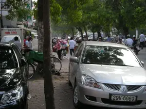 Đây là một cảnh đường phố nhộn nhịp ở Hà Nội. Vỉa hè được lát gạch và có nhiều cây xanh lớn. Nhiều người đang đi lại trên vỉa hè và di chuyển bằng xe máy dưới lòng đường. Một số người đi xe máy có đội mũ bảo hiểm. Ở phía trước, một chiếc ô tô màu bạc và một chiếc ô tô màu đen đang đỗ trên vỉa hè. Gần đó, một chiếc xe đạp được phủ tấm bạt màu xanh lá cây dựng cạnh gốc cây. Một người phụ nữ đang đẩy một chiếc xe đạp khác chở đầy hàng hóa cồng kềnh, cho thấy hoạt động buôn bán trên đường phố. Ở phía sau, có một chiếc xe tải nhỏ màu trắng với dòng chữ "ĐIỆN TỬ HẢI TÂU" trên thùng xe. Có nhiều tòa nhà và các biển hiệu khác ở hậu cảnh nhưng không thể đọc rõ.。（日本語訳）。これはハノイの賑やかな街頭の風景です。歩道はタイルで舗装されており、大きな緑の木がたくさんあります。多くの人が歩道を歩き、車道ではバイクで移動しています。バイクに乗っている人の中にはヘルメットをかぶっている人もいます。前方には、銀色の車と黒い車が歩道に駐車しています。その近くでは、緑色のシートで覆われた自転車が木の根元に立てかけられています。一人の女性が、かさばる商品をたくさん積んだ別の自転車を押しており、路上での商売の様子がうかがえます。後方には、荷台に「ĐIỆN TỬ HẢI TÂU」と書かれた白い小型トラックがあります。