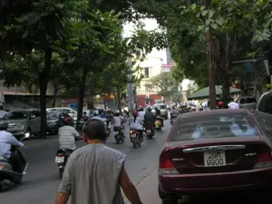 Đây là khung cảnh trên một con phố ở Hà Nội. Con đường có mật độ giao thông đông đúc với rất nhiều người đang điều khiển xe máy. Nhiều người trong số họ đội mũ bảo hiểm. Ngoài xe máy, còn có một vài chiếc ô tô. Nổi bật ở phía trước bên phải là một chiếc ô tô màu đỏ. Ở tiền cảnh, một người đàn ông mặc áo ba lỗ màu xám đang đi bộ trên đường. Hai bên đường được trồng nhiều cây xanh, tạo bóng mát. Phía sau là những tòa nhà cao tầng và các cửa hàng ven đường. Có thể thấy một số biển hiệu nhưng chữ viết trên đó không thể đọc được do mờ.。（日本語訳）。これはハノイのある通りの風景です。道路は交通量が多く、たくさんの人がバイクを運転しています。彼らの多くはヘルメットをかぶっています。バイクの他に、数台の自動車もあります。右手前に目立っているのは、一台の赤い自動車です。手前には、灰色のタンクトップを着た男性が道を歩いています。道の両側にはたくさんの緑の木が植えられており、日陰を作っています。後方には高層ビルや道端の店があります。いくつかの看板が見えますが、書かれている文字はぼやけていて読めません。
