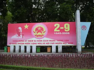 Bối cảnh là một không gian công cộng ngoài trời, có thể là công viên hoặc ven đường, với nhiều cây xanh ở phía sau. Một tấm pano tuyên truyền lớn màu đỏ được dựng lên trên một bệ bê tông. Trên pano có các nội dung được ghi lại như sau: Phía trên bên trái là chân dung Chủ tịch Hồ Chí Minh, ở giữa là Quốc huy Việt Nam với dòng chữ 'VIỆT NAM' bên dưới, và bên phải là dòng số '2-9' và '1945-2008'. Dòng chữ lớn ở dưới cùng là 'CHÀO MỪNG KỶ NIỆM 63 NĂM CÁCH MẠNG THÁNG TÁM VÀ QUỐC KHÁNH NƯỚC CỘNG HÒA XÃ HỘI CHỦ NGHĨA VIỆT NAM'. Hoạt động được thể hiện là việc trưng bày pano để kỷ niệm một sự kiện quốc gia. Phía trước là một luống hoa màu đỏ. Bên dưới pano có một vài thùng rác công cộng. Bên phải tấm pano màu đỏ là một biểu ngữ màu xanh lam có in biểu tượng của Hà Nội (Khuê Văn Các).。（日本語訳）。背景は屋外の公共スペースで、公園か道端の可能性があり、後ろには多くの緑の木々があります。赤い大きな宣伝パネルがコンクリートの台座の上に設置されています。パネルには以下の内容が記されています。左上にはホーチミン主席の肖像画、中央には下に「VIỆT NAM」の文字が入ったベトナムの国章、そして右側には「2-9」と「1945-2008」という数字の列があります。一番下の大きな文字は「ベトナム社会主義共和国の八月革命及び国慶節63周年記念を祝う」です。表現されている活動は、国家的な出来事を記念するためのパネルの展示です。手前には赤い花壇があります。パネルの下にはいくつかの公共のゴミ箱があります。赤いパネルの右側には、ハノイのシンボル（奎文閣）が印刷された青い横断幕があります。
