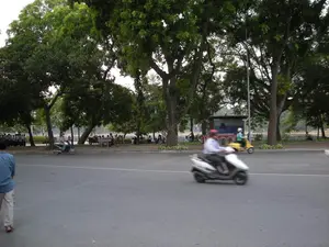 Bức ảnh chụp một khung cảnh đường phố ở Hà Nội, có lẽ là khu vực ven hồ. Con đường được trải nhựa, hai bên là vỉa hè rợp bóng cây cổ thụ xanh tốt. Trên đường, có vài chiếc xe máy (xe tay ga) đang di chuyển; nổi bật là một chiếc xe máy bị mờ do tốc độ, do một người đàn ông đội mũ bảo hiểm đỏ điều khiển. Trên vỉa hè, rất đông người đang tụ tập. Một số ngồi nghỉ trên ghế đá, số khác đứng nói chuyện bên cạnh những chiếc xe máy và xe đạp dựng ở đó. Trong hậu cảnh, có một công trình giống như nhà chờ xe buýt với các tấm áp phích không thể đọc được chữ. Phía sau hàng cây có thể nhìn thấy một phần của một mặt hồ. Một người đàn ông mặc áo xanh đang đi bộ ở phía bên trái của bức ảnh.。（日本語訳）。この写真はハノイのある街の風景を撮影したもので、おそらく湖畔のエリアでしょう。道路はアスファルトで舗装されており、両側には緑豊かな古い木々の木陰に覆われた歩道があります。道路上では、数台のバイク（スクーター）が走行しています。目立っているのは、赤いヘルメットをかぶった男性が運転する、速度のためにぼやけている一台のバイクです。歩道には、多くの人々が集まっています。石のベンチに座って休んでいる人もいれば、そこに停めてあるバイクや自転車のそばで立ち話をしている人もいます。背景には、文字が読み取れないポスターが貼られた、バスの待合所のような建物があります。木々の列の後ろに、湖面の一部が見えます。青いシャツを着た男性が、写真の左側を歩いています。
