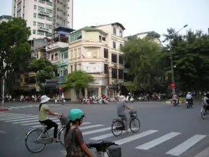 Bối cảnh là một ngã tư đường phố đông đúc ở một thành phố, có khả năng là Hà Nội, với sự pha trộn giữa kiến trúc cũ và mới. Ở hậu cảnh, một tòa nhà cao tầng hiện đại tương phản với những ngôi nhà ống nhiều tầng, san sát nhau, một kiểu kiến trúc đặc trưng của Việt Nam. Tầng trệt của các tòa nhà là các cửa hàng. Có một biển hiệu trên tòa nhà ở góc phố nhưng chữ viết bị mờ, không thể đọc rõ. Hoạt động chính là cảnh giao thông nhộn nhịp của người dân. Nhiều người đang di chuyển bằng xe máy và xe đạp. Ở tiền cảnh, có hai người phụ nữ đang đi trên một chiếc xe đạp qua vạch kẻ đường dành cho người đi bộ. Một người phụ nữ khác, lớn tuổi hơn và đội nón rộng vành, cũng đang đi xe đạp. Một người ngồi trên xe máy ở phía trước đội mũ bảo hiểm màu xanh ngọc. Nhiều xe máy khác đang lưu thông hoặc dừng chờ trên đường. Có một biển báo giao thông "Cấm đi ngược chiều" màu đỏ có thể nhìn thấy. Cây xanh được trồng dọc theo vỉa hè, tạo nên một khung cảnh đường phố điển hình.。（日本語訳）。背景は、ある都市、おそらくハノイの、新旧の建築が混在する賑やかな交差点です。背景には、近代的な高層ビルが、ベトナム特有の建築様式である、密集した多層階のチューブハウスと対照をなしています。建物の1階は店舗になっています。角の建物には看板がありますが、文字がぼやけていてはっきりと読むことはできません。主な活動は、人々の賑やかな交通風景です。多くの人々がバイクや自転車で移動しています。前景には、2人の女性が自転車に乗って横断歩道を渡っています。別の、より年配でつばの広い帽子をかぶった女性も自転車に乗っています。前方にいるバイクに乗った人は、青緑色のヘルメットをかぶっています。他にも多くのバイクが道路を走行しているか、停止して待っています。赤い「逆走禁止」の交通標識が見えます。歩道に沿って緑の木々が植えられており、典型的な街の風景を作り出しています。