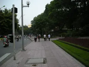 Đây là một cảnh quan đô thị tại Hà Nội. Hình ảnh cho thấy một con đường nhựa ở bên trái với nhiều người đang đi xe máy và một chiếc ô tô. Một số người đi xe máy đội mũ bảo hiểm. Ở giữa là một vỉa hè rộng được lát gạch, nơi một số người đang đi bộ. Dọc vỉa hè có một cột đèn đường lớn và một cột tín hiệu giao thông. Bên phải là một khu vực công viên hoặc dải cây xanh với rất nhiều cây lớn, rậm rạp, cùng với các luống cây cảnh và cỏ được cắt tỉa gọn gàng. Bầu trời có vẻ như là vào lúc hoàng hôn hoặc sáng sớm. Ở phía xa có thể nhìn thấy một tòa nhà. Không có văn bản nào có thể đọc được.。（日本語訳）。これはハノイの都市景観です。画像は、左側にアスファルトの道路があり、多くの人々がバイクに乗り、1台の車が写っていることを示しています。バイクに乗っている人の中にはヘルメットをかぶっている人もいます。中央にはタイル張りの広い歩道があり、数人が歩いています。歩道に沿って、大きな街灯柱と信号機の柱があります。右側は公園または緑地帯で、多くの大きくて生い茂った木々があり、きれいに手入れされた植え込みや芝生もあります。空は夕暮れ時か早朝のようです。遠くに建物が見えます。読み取れるテキストはありません。
