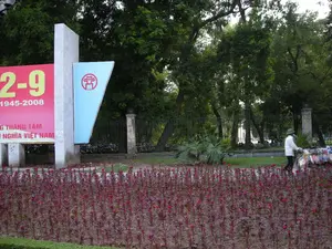 Bối cảnh là một công viên hoặc vườn hoa công cộng ở Hà Nội. Ở phía trước là một luống hoa màu đỏ tía, có thể là hoa mào gà. Ở phía sau là nhiều cây xanh lớn, bãi cỏ và một hàng rào sắt.。Bên trái có một tấm pano lớn màu đỏ và một lá cờ trang trí. Trên tấm pano có các dòng chữ sau:。- Số lớn màu vàng: "2-9"。- Dãy số bên dưới: "1945-2008"。- Dòng chữ tiếng Việt (một phần bị che khuất): "...NG THÁNG TÁM" và "...NGHĨA VIỆT NAM", có thể đọc đầy đủ là "CHÀO MỪNG CÁCH MẠNG THÁNG TÁM VÀ QUỐC KHÁNH NƯỚC CỘNG HÒA XÃ HỘI CHỦ NGHĨA VIỆT NAM". Tấm pano này được dựng lên để kỷ niệm Quốc khánh Việt Nam ngày 2 tháng 9.。Bên cạnh tấm pano là một lá cờ hình tam giác màu xanh nhạt có in biểu trưng của Hà Nội (hình ảnh Khuê Văn Các).。Ở phía bên phải, một người đội nón lá, mặc áo trắng đang đi bộ và đẩy một chiếc xe bán hàng rong chứa nhiều loại hàng hóa.。（日本語訳）。背景はハノイにある公園または公共の花園です。手前には赤紫色の花壇があり、おそらくケイトウの花でしょう。後方には多くの大きな緑の木々、芝生、そして鉄の柵があります。左側には大きな赤色の看板と一つの装飾用の旗があります。看板には以下の文字列があります：。- 黄色の大きな数字：「2-9」。- その下にある数字の列：「1945-2008」。- ベトナム語の文字列（一部が隠れている）：「...NG THÁNG TÁM」と「...NGHĨA VIỆT NAM」。全文はおそらく「CHÀO MỪNG CÁCH MẠNG THÁNG TÁM VÀ QUỐC KHÁNH NƯỚC CỘNG HÒA XÃ HỘI CHỦ NGHĨA VIỆT NAM」（八月革命とベトナム社会主義共和国建国記念日を祝う）と読めます。この看板は9月2日のベトナム建国記念日を記念して設置されたものです。看板の隣には、ハノイのシンボル（奎文閣の絵）が印刷された水色の三角形の旗があります。右側では、ノンラーをかぶり白い服を着た一人の人物が、様々な商品を積んだ移動販売のカートを押しながら歩いています。