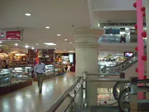 Bối cảnh là bên trong một trung tâm thương mại hoặc siêu thị nhiều tầng. Có một số biển báo có thể đọc được. Một biển báo màu đỏ và trắng treo trên trần nhà ghi 'LỐI THOÁT HIỂM' và 'EXIT'. Ở tầng trên, có một biển hiệu khác ghi 'SIÊU THỊ VI...', các chữ còn lại bị che khuất. Một biển hiệu khác ở bên trái có chữ 'HÀNG B...' nhưng không rõ phần còn lại.。Hình ảnh mô tả hoạt động mua sắm và đi lại trong không gian thương mại. Một người đàn ông mặc áo sơ mi sáng màu và quần tây đang đi bộ trong hành lang. Nhiều người khác cũng có mặt, một số đang xem hàng ở các gian hàng bên trái, một số đang di chuyển bằng thang cuốn ở bên phải. Các gian hàng bên trái trưng bày các sản phẩm trong tủ kính, có vẻ là đồ lưu niệm, tranh ảnh nghệ thuật và có thể là trang sức. Có một cây cột lớn, màu kem ở giữa. Tòa nhà có ít nhất hai tầng, được kết nối bằng thang cuốn. Một cây cột trang trí màu hồng được gắn các quả cầu màu đỏ, có thể là đèn lồng cho một dịp lễ hội.。（日本語訳）。背景は、ある商業施設または多層階のスーパーマーケットの中です。読むことができる看板がいくつかあります。天井から吊るされた赤と白の看板には、「LỐI THOÁT HIỂM」と「EXIT」と書かれています。上の階には、「SIÊU THỊ VI...」と書かれた別の看板があり、残りの文字は隠れています。左側にある別の看板には「HÀNG B...」という文字がありますが、残りの部分ははっきりしません。画像は、商業空間での買い物や行き来の様子を描写しています。明るい色のシャツとズボンを履いた男性が廊下を歩いています。他にも多くの人々がおり、何人かは左側の売店で商品を見ており、何人かは右側でエスカレーターを使って移動しています。左側の売店はガラスケースの中に商品を展示しており、お土産、芸術的な絵画や写真、そしておそらく宝飾品のようです。中央にはクリーム色の大きな柱があります。建物は少なくとも2階建てで、エスカレーターで結ばれています。ピンク色の装飾的な柱には赤い球が取り付けられており、これは祭事のための提灯かもしれません。