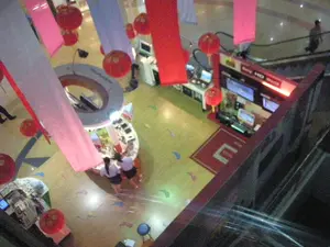 Bức ảnh được chụp từ trên cao xuống, cho thấy khung cảnh bên trong một trung tâm thương mại hoặc trung tâm điện máy. Không gian được trang trí cho một sự kiện hoặc lễ hội, với nhiều đèn lồng đỏ và các dải băng vải nhiều màu sắc (đỏ, trắng, hồng) treo từ trần nhà. Ở trung tâm có một quầy hàng hình tròn, nơi hai người phụ nữ mặc đồng phục (áo trắng, váy sẫm màu) đang đứng, có thể là nhân viên. Bên phải là một quầy hàng khác chuyên về đồ điện tử, trưng bày nhiều màn hình TV hoặc máy tính. Trên quầy này có một biển hiệu phát sáng màu đỏ với chữ "HD" có thể đọc được, các chữ còn lại bị mờ. Phía trước quầy hàng này là một tấm thảm đỏ có một ký tự hoặc logo lớn màu trắng, trông giống chữ "E". Ở phía sau, có thể thấy một người đang đi trên thang cuốn. Sàn nhà có màu sáng và được trang trí bằng những hình dán nhỏ nhiều màu sắc.。（日本語訳）。写真は上から撮影されたもので、ショッピングモールまたは家電量販店の内部の光景を写しています。空間はイベントか祭りのために装飾されており、たくさんの赤い提灯と多色（赤、白、ピンク）の布製リボンが天井から吊るされています。中央には円形のカウンターがあり、そこには制服（白いシャツ、暗い色のスカート）を着た2人の女性が立っており、従業員である可能性があります。右側には電子機器を専門とする別のカウンターがあり、多くのテレビまたはコンピュータの画面が展示されています。このカウンターの上には赤く光る看板があり、「HD」という文字が読み取れますが、残りの文字はぼやけています。後方では、エスカレーターに乗っている人が見えます。床は明るい色で、小さくカラフルなステッカーで装飾されています。