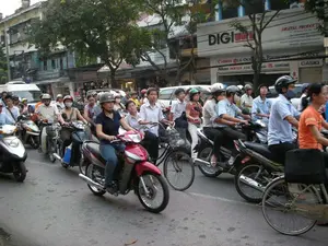 Bức ảnh ghi lại cảnh tượng giao thông đông đúc trên một con phố ở Hà Nội. Dòng phương tiện, chủ yếu là xe máy, đang dừng lại hoặc di chuyển rất chậm, cho thấy khả năng đang có ùn tắc giao thông.。**Chữ viết được ghi lại:**。Trên biển hiệu của một tòa nhà lớn, có các dòng chữ:。*   "DIGIworld"。*   "DIGITAL PRODUCTS"。*   Bên dưới là danh sách các thương hiệu điện tử: "Canon", "LG", "OLYMPUS" (OLY), "Panasonic", "SONY", "Nikon", "PENTAX", "JVC" (JUNE), "Apple" (ple), "JBL", "SAMSUNG", "CASIO".。*   Có thể nhận ra một phần địa chỉ là "Hang Ba".。**Mô tả cảnh vật và hoạt động:**。*   **Loại địa điểm:** Một con phố đô thị sầm uất, hai bên là các tòa nhà cao tầng liền kề và cây xanh.。*   **Hoạt động cụ thể:** Mọi người đang tham gia giao thông. Các phương tiện đang dừng lại thành hàng dài, có thể là đang chờ đèn tín hiệu hoặc do mật độ xe quá đông vào giờ cao điểm.。*   **Vật thể:**。*   **Phương tiện:** Rất nhiều xe máy (xe gắn máy) đủ loại. Có một chiếc xe đạp ở góc dưới bên phải và một chiếc xe van màu trắng ở phía sau.。*   **Con người:** Người dân ở nhiều độ tuổi đang điều khiển xe máy. Hầu hết mọi người đều đội mũ bảo hiểm. Một số người đeo khẩu trang.。*   **Khác:** Các tòa nhà được sử dụng làm nhà ở và cửa hàng kinh doanh, nổi bật là cửa hàng bán đồ kỹ thuật số.。（日本語訳）。写真は、ハノイのある通りでの混雑した交通の光景を記録しています。主にバイクからなる車両の流れは、停止しているか、非常にゆっくりと移動しており、交通渋滞が発生している可能性を示しています。**記録されている文字：**。大きな建物の看板には、以下の文字があります：。*   "DIGIworld"。*   "DIGITAL PRODUCTS"。*   その下には電子機器ブランドのリストがあります：「Canon」、「LG」、「OLYMPUS」（OLY）、「Panasonic」、「SONY」、「Nikon」、「PENTAX」、「JVC」（JUNE）、「Apple」（ple）、「JBL」、「SAMSUNG」、「CASIO」。*   住所の一部が「Hang Ba」であることが認識できます。**風景と活動の説明：**。*   **場所の種類：** 賑やかな都市の通りで、両側には隣接する高層ビルと緑の木々があります。*   **具体的な活動：** 人々が交通に参加しています。車両は長い列を作って停止しており、信号を待っているか、あるいはラッシュアワーで交通量が多すぎるためかもしれません。*   **物体：**。*   **車両：** 非常に多くの様々な種類のバイク（原動機付自転車）。右下隅に自転車が1台、後方に白いバンが1台あります。*   **人物：** 様々な年齢の人々がバイクを運転しています。ほとんどの人がヘルメットを着用しています。マスクを着用している人もいます。*   **その他：** 建物は住居や店舗として使用されており、特に目立っているのはデジタル製品を販売する店です。