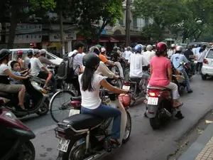 Bức ảnh ghi lại cảnh giao thông đông đúc trên một con đường đô thị, có thể là ở Hà Nội. Dòng người và phương tiện, chủ yếu là xe máy, đang nối đuôi nhau trên đường. Hầu hết mọi người tham gia giao thông đều đội mũ bảo hiểm. Có thể thấy nhiều loại người khác nhau, từ người đi làm, phụ nữ chở con nhỏ ngồi phía trước, cho đến học sinh đeo ba lô. Hai bên đường là những tòa nhà nhiều tầng với các cửa hàng ở tầng trệt và cây xanh. Một số biển hiệu có thể được nhận dạng một phần, chẳng hạn như 'ELIT' và 'BLACK F...'. Các biển hiệu khác thì không thể đọc rõ. Đây là một cảnh tượng điển hình về giao thông vào giờ cao điểm tại một thành phố lớn của Việt Nam.。（日本語訳）。写真は、おそらくハノイの、都市部の道路における混雑した交通風景を記録しています。人と乗り物、主にバイクの流れが、道路で次々と連なっています。交通に参加しているほとんどの人がヘルメットを着用しています。通勤者、前に小さな子供を乗せた女性から、バックパックを背負った学生まで、さまざまな種類の人々が見られます。道路の両側には、1階に店舗が入った多層階の建物と緑の木々があります。いくつかの看板は、「ELIT」や「BLACK F...」のように、部分的に識別できます。他の看板ははっきりと読み取れません。これは、ベトナムの大都市におけるラッシュアワーの交通の典型的な光景です。