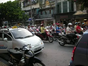 Các thông tin bằng chữ có thể nhận dạng bao gồm: biển hiệu "TAXI" màu vàng trên nóc một chiếc ô tô; logo và dòng chữ "THÀNH NAM" cùng số điện thoại "...91.91" trên cửa xe taxi này. Ngoài ra còn có biển hiệu cửa hàng ghi "DIGI..." nhưng không rõ phần còn lại.。Bối cảnh là một con phố đông đúc tại một khu đô thị, có thể là Hà Nội. Hoạt động chính đang diễn ra là giao thông vào giờ cao điểm, với một lượng lớn xe máy đang lưu thông trên đường. Mọi người điều khiển xe máy đều đội mũ bảo hiểm. Nổi bật trong ảnh là một chiếc ô tô nhỏ màu trắng đang hoạt động như một chiếc taxi, xen lẫn giữa dòng xe máy. Các vật thể khác bao gồm nhiều tòa nhà kiểu nhà ống san sát nhau, với các cửa hàng ở tầng trệt, và cây xanh ở bên lề đường.。（日本語訳）。認識可能な文字情報には、ある自動車の屋根にある黄色の「TAXI」の看板、このタクシーのドアにある「THÀNH NAM」のロゴと文字、および電話番号「...91.91」が含まれます。その他に、「DIGI...」と書かれた店の看板もありますが、残りの部分は不明です。背景は、ある都市部の混雑した通りで、おそらくハノイです。行われている主な活動はラッシュアワーの交通で、多数のバイクが道路を走行しています。バイクを運転している人々は皆ヘルメットを着用しています。写真の中で目立っているのは、タクシーとして営業している一台の白い小型車で、バイクの流れに混じっています。その他の物体には、1階に店舗がある密集して建つ多くのチューブハウス（縦長の家）スタイルの建物、そして道端の緑の木々が含まれます。