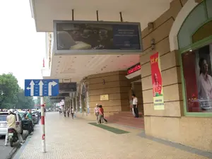 Bối cảnh là vỉa hè trước một tòa nhà lớn, hiện đại trên một con phố ở Hà Nội.。【Thông tin chữ viết】。*   Màn hình quảng cáo lớn treo dưới mái hiên có nội dung: "FORD ESCAPE MỚI KHÁC BIỆT TỪ MỘT PHONG CÁCH SUV ĐÍCH THỰC" cùng với logo của hãng xe "Ford".。*   Biển báo giao thông màu xanh chỉ dẫn hướng đi trên các làn đường: làn ngoài cùng bên trái cho phép rẽ trái, hai làn giữa đi thẳng, và làn ngoài cùng bên phải cho phép rẽ phải.。*   Băng rôn tuyên truyền màu đỏ được treo trên cột của tòa nhà, có hình ngôi sao vàng, dòng chữ "2-9" và hình ảnh Chủ tịch Hồ Chí Minh, có thể là để kỷ niệm ngày Quốc khánh.。*   Bảng điện tử màu đỏ ở lối vào tòa nhà có dòng chữ "...TOÀN THỂ CB...", phần chữ còn lại không nhìn rõ.。【Miêu tả cảnh vật và hoạt động】。*   **Địa điểm:** Đây là vỉa hè rộng rãi trước một tòa nhà có kiến trúc hiện đại, tường ốp đá màu be. Lối đi có mái che. Bên lề đường là một con phố đông đúc với nhiều cây xanh ở phía xa.。*   **Hoạt động:** Nhiều người đang đi bộ trên vỉa hè. Một người đàn ông mặc áo sơ mi trắng đang đứng trên bậc tam cấp ở lối vào chính của tòa nhà. Một nhóm người khác đang đi về phía người xem. Dưới lòng đường, một người đội mũ bảo hiểm đang ngồi trên chiếc xe máy đang đỗ. Có nhiều ô tô và xe máy khác đang dừng hoặc di chuyển trên đường. Hoạt động chung là cảnh sinh hoạt đô thị thường ngày.。*   **Vật thể:** Tòa nhà, vỉa hè, biển báo giao thông, màn hình quảng cáo, băng rôn, xe máy, ô tô, cây cối và người đi đường.。（日本語訳）。背景は、ハノイのある通りにある、大きくて近代的なビルの前の歩道です。【文字情報】。*   軒下に吊るされた大きな広告スクリーンには、自動車メーカー「フォード」のロゴと共に、「FORD ESCAPE MỚI KHÁC BIỆT TỪ MỘT PHONG CÁCH SUV ĐÍCH THỰC」（新しいフォード・エスケープ、本物のSUVスタイルが生み出す違い）という内容があります。*   青い交通標識が各車線の進行方向を示しています。一番左の車線は左折可、中央の2車線は直進、そして一番右の車線は右折可です。*   ビルの柱に掲げられた赤い宣伝横断幕には、黄色い星、「2-9」という文字、そしてホー・チ・ミン主席の肖像画があり、建国記念日を祝うためのものと思われます。*   ビルの入り口にある赤い電光掲示板には「...TOÀN THỂ CB...」という文字があり、残りの文字ははっきりと見えません。【風景と活動の描写】。*   **場所:** これは、ベージュ色の石で覆われた壁を持つ近代的な建築のビルの前にある、広々とした歩道です。通路には屋根があります。歩道の脇には賑やかな通りがあり、遠くには緑の木々が多く見えます。*   **活動:** 多くの人が歩道を歩いています。白いシャツを着た男性が、ビルの正面玄関の階段に立っています。別のグループが鑑賞者の方へ向かって歩いています。車道では、ヘルメットをかぶった人が停車中のバイクに座っています。他にも多くの車やバイクが道路で停車または走行しています。全体的な活動は、日常的な都市の生活風景です。*   **物体:** ビル、歩道、交通標識、広告スクリーン、横断幕、バイク、自動車、木々、そして通行人。