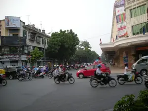 Bức ảnh chụp một ngã tư đường phố đông đúc tại Hà Nội. Khung cảnh cho thấy dòng phương tiện giao thông tấp nập, chủ yếu là xe máy, đang di chuyển trên đường. Nhiều người điều khiển xe máy đội mũ bảo hiểm. Một chiếc ô tô màu đỏ đang ở giữa ngã tư, cùng với các loại xe ô tô và xe máy khác. Hai bên đường là các tòa nhà. Bên trái là một dãy nhà phố kiểu cũ, nhiều tầng. Bên phải là một tòa nhà lớn, có kiến trúc hiện đại hơn, trông giống một trung tâm thương mại. Có một vài người đi bộ trên vỉa hè. Cây xanh được trồng dọc theo đường phố. Các dòng chữ có thể nhận dạng được bao gồm: trên nóc tòa nhà bên trái có một biển quảng cáo lớn với nội dung '500 GIỜ TẬN HƯỞNG CHO LY BIA...', phía dưới là biển quảng cáo 'RADO' và biển hiệu cửa hàng 'California DVD KARAOKE VI TÍNH'. Đèn tín hiệu giao thông đang hiển thị số '15'. Trên tòa nhà bên phải có một tấm áp phích quảng cáo với chữ 'Caryn'.。（日本語訳）。ハノイの混雑した交差点を撮影した写真です。多くのバイク運転手はヘルメットを着用しています。赤い車が1台、他の様々な車やバイクと共に交差点の真ん中にいます。道路の両側には建物があります。左側には古い様式の多層階のタウンハウスが並んでいます。右側には、より近代的な建築の大きな建物があり、ショッピングセンターのように見えます。歩道には数人の歩行者がいます。街路樹が通りに沿って植えられています。認識できる文字には、左側の建物の屋上にある「500 GIỜ TẬN HƯỞNG CHO LY BIA...」（ビール一杯を楽しむ500時間...）という内容の大きな広告看板、その下には「RADO」の広告看板と「California DVD KARAOKE VI TÍNH」という店の看板が含まれます。交通信号機は「15」という数字を表示しています。右側の建物には「Caryn」という文字の広告ポスターがあります。