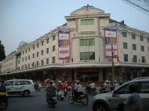 Bức ảnh chụp cảnh đường phố đông đúc trước một tòa nhà lớn ở Hà Nội vào lúc chiều tối.。**Chữ viết được ghi lại:**。*   Trên đỉnh tòa nhà có dòng chữ: "TRUNG TÂM THƯƠNG MẠI TRÀNG TIỀN".。*   Hai biểu ngữ lớn treo trên tòa nhà có logo "Great Eastern" và các dòng chữ quảng cáo như "CÙNG BẠN ĐI ĐẾN TƯƠNG LAI", "PHÒNG TỎA NÉT VIỆT NAM", "Singapore và Malaysia".。*   Trên thân một chiếc xe taxi màu trắng có chữ "SMARTDOOR" và số điện thoại "38 53 53 53".。**Mô tả cảnh vật và hoạt động:**。*   **Loại địa điểm:** Đây là một con đường lớn ở khu vực trung tâm thành phố, phía trước Trung tâm Thương mại Tràng Tiền, một tòa nhà có kiến trúc kiểu Pháp.。*   **Hoạt động cụ thể:** Dòng người và phương tiện đang tham gia giao thông tấp nập. Mọi người đang lái xe máy và ô tô trên đường. Một người đi xe máy đang chở một chùm bóng bay nhiều màu sắc. Đây là cảnh sinh hoạt đô thị điển hình.。*   **Các vật thể trong ảnh:**。*   **Tòa nhà:** Trung tâm Thương mại Tràng Tiền, một tòa nhà nhiều tầng màu kem, được trang trí bằng dây đèn và các lá cờ nhỏ.。*   **Phương tiện:** Rất nhiều xe máy (xe máy), một chiếc taxi, và các loại ô tô con.。*   **Con người:** Nhiều người đang điều khiển phương tiện giao thông, hầu hết đều đội mũ bảo hiểm.。*   **Khác:** Các biểu ngữ quảng cáo, cột đèn đường, dây điện, và đèn giao thông.。（日本語訳）。ハノイの大きな建物の前にある、夕暮れ時の混雑した通りの光景を撮影した写真です。**記録されている文字：**。*   建物の最上部には「TRUNG TÂM THƯƠNG MẠI TRÀNG TIỀN」（チャンティエン・ショッピングセンター）という文字があります。*   建物に掲げられた2つの大きな横断幕には、「Great Eastern」のロゴと、「CÙNG BẠN ĐI ĐẾN TƯƠNG LAI」（あなたと共に未来へ）、「PHÒNG TỎA NÉT VIỆT NAM」（ベトナムの美しさを解き放つ）、「Singapore và Malaysia」（シンガポールとマレーシア）といった広告文があります。*   白いタクシーの車体には「SMARTDOOR」という文字と電話番号「38 53 53 53」があります。**風景と活動の説明：**。*   **場所の種類：** これは市の中心部にある大きな通りで、フランス様式の建築物であるチャンティエン・ショッピングセンターの前にあります。*   **具体的な活動：** 人と車両の流れで交通が混雑しています。人々は道路でバイクや車を運転しています。バイクに乗った一人の人が、色とりどりの風船の束を運んでいます。これは典型的な都市の生活風景です。*   **写真の中の物体：**。*   **建物：** チャンティエン・ショッピングセンター、クリーム色の多層階の建物で、電飾や小さな旗で飾られています。*   **乗り物：** 非常に多くのバイク（xe máy）、1台のタクシー、そして様々な種類の乗用車。*   **人々：** 多くの人々が乗り物を運転しており、ほとんどがヘルメットを着用しています。*   **その他：** 広告の横断幕、街灯、電線、そして信号機。