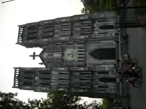 Bức ảnh chụp Nhà thờ Lớn Hà Nội, một công trình kiến trúc Gothic cổ kính. Nhà thờ có màu xám, với hai tháp chuông cao đối xứng. Bức tường có vẻ cũ và nhuốm màu thời gian. Ở chính giữa mặt tiền, phía trên cửa ra vào chính, có một chiếc đồng hồ lớn và một biểu tượng hình tròn có hình vẽ tôn giáo. Trên đỉnh cao nhất là một cây thánh giá. Phía trước nhà thờ là một con đường, nơi có một số người dân và du khách đang đứng và đi lại. Một số người đang ngồi trên hoặc đứng cạnh xe máy đang đỗ. Cảnh vật cho thấy một ngày sinh hoạt bình thường tại một địa danh nổi tiếng của thành phố. Xung quanh có cây xanh và hàng rào sắt.。（日本語訳）。ハノイ大教会を撮影した写真、古いゴシック建築の建造物です。教会は灰色で、左右対称の高い鐘楼が二つあります。壁は古く見え、時の色を帯びています。ファサードの中央、正面玄関の上には、大きな時計と宗教的な絵が描かれた円形のシンボルがあります。一番高い頂上には十字架があります。教会の前には道があり、そこには何人かの地元の人々と観光客が立ったり歩いたりしています。何人かの人々は、駐車しているバイクに座ったり、その隣に立ったりしています。この光景は、市の有名な名所での日常の活動の一日を示しています。周りには緑の木々と鉄の柵があります。