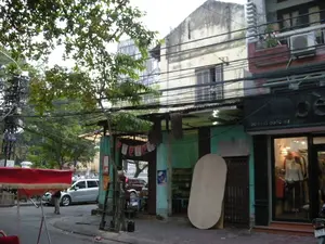 Bản ghi văn bản:。- Biển tên đường màu xanh có chữ: AU TRIEU.。- Trên biển hiệu của cửa hàng bên phải có ghi địa chỉ: ĐC: 58 LÝ QUỐC SƯ.。Mô tả cảnh quan và hoạt động:。Đây là khung cảnh trên một con phố ở Hà Nội. Phía xa, qua những tán cây, có thể nhìn thấy một phần của một công trình kiến trúc lớn kiểu Gothic, có khả năng là Nhà thờ Lớn Hà Nội.。- Địa điểm: Một con phố với những ngôi nhà ống san sát. Một số ngôi nhà trông cũ kỹ với lớp sơn đã bạc màu, trong khi một ngôi nhà khác ở bên phải là một cửa hàng thời trang có mặt tiền màu đen và cửa kính sáng sủa.。- Hoạt động: Cảnh sinh hoạt đường phố thường ngày. Một người đàn ông, có lẽ là người lái xích lô, đang đứng bên cạnh chiếc xe xích lô có mái che màu đỏ của mình. Một cửa hàng quần áo đang mở cửa, bên trong trưng bày ma-nơ-canh. Có ô tô và xe máy đang đỗ trên đường.。- Các vật thể nổi bật: Một mạng lưới dây điện và cáp viễn thông chằng chịt phía trên đường phố, một đặc trưng của các khu đô thị cũ ở Việt Nam. Một cây xanh lớn ở phía trước. Một chiếc xe xích lô, một chiếc ô tô màu bạc, và một phần của một chiếc xe máy. Dựa vào tường là một tấm ván lớn hình bầu dục và một chiếc thang gỗ. Một vài lá cờ nhỏ màu đỏ và trắng được treo phía trước một ngôi nhà.。（日本語訳）。テキスト記録：。- 青い道路標識に「AU TRIEU」の文字。- 右側の店の看板に住所が記載されている：住所：58 LY QUOC SU。風景と活動の説明：。これはハノイのある通りでの光景です。遠く、木々の向こうに、ゴシック様式の大きな建築物の一部が見え、それはハノイ大教会である可能性があります。- 場所：間口が狭く奥行きのある家々が密集する通り。いくつかの家は塗装が色褪せて古びて見えますが、右側にある別の家は黒いファサードと明るいガラス扉を持つファッション店です。- 活動：日常的な通りの生活風景。一人の男性、おそらくシクロの運転手が、赤い幌のついた自分のシクロの隣に立っています。一軒の衣料品店が開店しており、中にはマネキンが展示されています。道路には自動車とバイクが駐車されています。- 目立つ物体：通りの上には、ベトナムの古い都市部の特徴である、複雑に絡み合った電線と通信ケーブルの網。前面に大きな緑の木。一台のシクロ、一台の銀色の自動車、そして一台のバイクの一部。壁には大きな楕円形の板と木製のはしごが立てかけられています。家の前に、いくつかの小さな赤と白の旗が掲げられています。