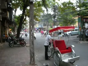 Bức ảnh chụp một khung cảnh trên một con phố ở Hà Nội. Toàn cảnh khá mờ. Ở phía trước, một người đàn ông mặc áo sơ mi trắng đang đứng bên cạnh một chiếc xe xích lô chở khách. Chiếc xích lô có phần ghế ngồi cho khách được bọc nệm vải màu đỏ và khung xe bằng kim loại sáng bóng. Người đàn ông có vẻ là người lái xe đang chờ khách. Dọc vỉa hè có cây xanh, một cột điện bê tông và một vài chiếc xe máy đang đậu. Phía sau là đường phố với nhiều người và phương tiện qua lại, bao gồm cả người đi bộ và xe máy. Các tòa nhà liền kề nhau nằm dọc hai bên đường. Không có chữ viết nào có thể đọc được do ảnh bị mờ.。（日本語訳）。写真はハノイのある通りでの光景を写しています。全体的にかなりぼやけています。手前には、白いシャツを着た男性が客を乗せるシクロの隣に立っています。そのシクロは、客が座る座席部分が赤い布のクッションで覆われ、車体は光沢のある金属製です。その男性は客を待っている運転手のようです。歩道沿いには緑の木々、コンクリートの電柱、そして数台のバイクが停まっています。後ろには、歩行者やバイクを含む多くの人々や乗り物が行き交う通りがあります。通りの両側には建物が隣接して並んでいます。写真がぼやけているため、読み取れる文字はありません。