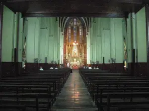 Bên trong một nhà thờ lớn mang phong cách kiến trúc Gothic. Không gian rộng lớn với trần nhà cao và các cột tường vững chãi. Hai bên là hai dãy ghế dài bằng gỗ sẫm màu dành cho giáo dân, được xếp thẳng hàng và còn rất nhiều chỗ trống. Một lối đi chính ở giữa trải dài từ phía người chụp ảnh đến khu vực cung thánh ở phía xa. Cung thánh được chiếu sáng, nổi bật với một bàn thờ và một bức tượng đặt trong hốc tường. Phía trên bàn thờ là cửa sổ kính màu đặc trưng của nhà thờ. Dọc các bức tường có gắn nhiều quạt điện. Một vài người đang ngồi trên các hàng ghế, dường như đang cầu nguyện hoặc nghỉ chân trong không gian tĩnh lặng và trang nghiêm của nhà thờ. Không có chữ viết nào có thể đọc được.。（日本語訳）。ゴシック建築様式の大聖堂の内部。高い天井と頑丈な柱壁を持つ広大な空間。両側には、信徒用の濃い色の木製長椅子が2列あり、まっすぐに並べられ、まだ空席がたくさんある。中央の主通路が、撮影者の側から遠くの聖域まで続いている。聖域は照らされており、祭壇と壁のくぼみに置かれた像が際立っている。祭壇の上には、教会特有のステンドグラスの窓がある。壁沿いには多くの扇風機が取り付けられている。数人が椅子に座っており、教会の静かで荘厳な空間で祈りを捧げているか、あるいは足を休めているようだ。読み取れる文字はない。