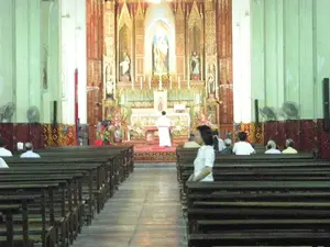 Bức ảnh chụp khung cảnh bên trong một nhà thờ Công giáo. Ở phía trước là khu vực cung thánh với một bàn thờ lớn, được trang trí công phu bằng gỗ chạm khắc và các chi tiết mạ vàng. Một linh mục mặc áo lễ màu trắng đang đứng trước bàn thờ, quay lưng về phía máy ảnh, dường như đang cử hành một buổi lễ. Phía sau bàn thờ có nhiều tượng thánh được đặt trong các hốc tường. Nhiều người dân (giáo dân) đang ngồi trên các hàng ghế gỗ dài, hướng về phía bàn thờ để tham dự buổi lễ. Các bức tường của nhà thờ có màu xanh nhạt, và có nhiều quạt điện được gắn trên tường. Lối đi ở giữa trống trải, dẫn thẳng đến bàn thờ.。（日本語訳）。カトリック教会内部の光景を撮影した写真です。前方には聖域があり、そこには木彫りや金メッキの細部で精巧に装飾された大きな祭壇があります。白い祭服を着た一人の司祭が祭壇の前に立ち、カメラに背を向けて、儀式を執り行っているようです。祭壇の後ろには、壁のくぼみに置かれた多くの聖像があります。多くの人々（信者）が、儀式に参加するために祭壇の方を向いて、長い木製のベンチに座っています。教会の壁は淡い青色で、壁には多くの扇風機が取り付けられています。中央の通路には何も置かれておらず、祭壇までまっすぐ続いています。