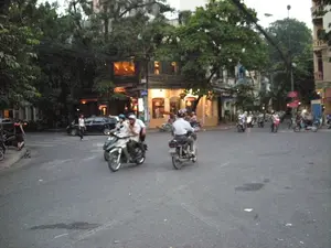 Bối cảnh là một góc phố hoặc ngã tư tại Hà Nội vào lúc chạng vạng tối. Đường phố tấp nập người và phương tiện qua lại, chủ yếu là xe máy. Ở phía trước, có hai chiếc xe máy đang di chuyển, một chiếc chở hai người và một chiếc chở một người. Tất cả những người đi xe máy đều đội mũ bảo hiểm. Ở phía sau, có nhiều xe máy khác đang lưu thông và một chiếc ô tô màu đen. Một người phụ nữ đang đi bộ qua đường. Hai bên đường là những tòa nhà nhiều tầng, tầng trệt có các cửa hàng đang sáng đèn. Rất nhiều cây xanh lớn, tán lá xum xuê bao phủ một phần con phố. Có thể thấy các cột đèn đường đã bật sáng và hệ thống dây điện chằng chịt phía trên. Có một số biển báo nhưng chữ viết trên đó quá mờ để có thể đọc được.。（日本語訳）。背景は、夕暮れ時のハノイの街角または交差点です。通りは人や乗り物で賑わっており、主にバイクです。手前には、2台のバイクが移動しており、1台は2人乗り、もう1台は1人乗りです。バイクに乗っている人は全員ヘルメットを着用しています。後方には、他の多くのバイクが走行しており、黒い自動車が1台あります。一人の女性が道を歩いて渡っています。道の両側には多層階の建物があり、1階には明かりの灯った店があります。たくさんの大きな緑の木があり、その青々とした葉が通り一部を覆っています。街灯が点灯し、頭上には電線が複雑に張り巡らされているのが見えます。いくつかの標識がありますが、そこに書かれている文字はぼやけていて読めません。