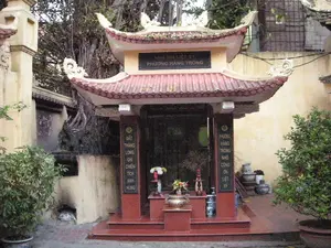 Đây là hình ảnh một nhà bia tưởng niệm liệt sỹ tại phường Hàng Trống, Hà Nội.。1. Chữ viết được ghi lại:。- Trên trán nhà bia: "NHÀ BIA LIỆT SỸ / PHƯỜNG HÀNG TRỐNG".。- Trên cột bên trái: "ĐẤT THĂNG LONG GHI CHIẾN TÍCH ANH HÙNG".。- Trên cột bên phải: "PHƯỜNG HÀNG TRỐNG NHỚ CÔNG ƠN LIỆT SỸ".。2. Bối cảnh và hoạt động:。- Loại địa điểm: Đây là một nhà bia tưởng niệm, một công trình kiến trúc nhỏ mang phong cách truyền thống với mái ngói đỏ uốn cong, nằm trong một khoảng sân nhỏ.。- Các vật thể trong ảnh: Bên trong nhà bia là một tấm bia đá lớn màu đen. Phía trước tấm bia là một bàn thờ màu đỏ, trên đó có đặt một lư hương bằng đồng, một bình hoa tươi và các vật phẩm thờ cúng khác. Hai bên lối vào là hai cột trụ màu đỏ có khắc câu đối. Xung quanh khu tưởng niệm có cây cối, đáng chú ý là một cây cổ thụ lớn ở phía sau và các chậu cây cảnh được đặt ở hai bên.。- Hoạt động: Mặc dù không có người trong ảnh, sự hiện diện của hoa tươi trên bàn thờ cho thấy hoạt động tưởng niệm và thờ cúng vẫn thường xuyên diễn ra tại đây, thể hiện lòng biết ơn của người dân đối với các anh hùng liệt sỹ.。（日本語訳）。これはハノイ市ハンチョン区にある烈士記念碑堂の写真です。1. 記されている文字：。- 碑堂の額：「烈士記念碑堂／ハンチョン区」。- 左側の柱：「タンロンの地は英雄の戦功を記す」。- 右側の柱：「ハンチョン区は烈士の恩に感謝する」。2. 背景と活動：。- 場所の種類：これは記念碑堂であり、小さな中庭に位置する、反り返った赤い瓦屋根を持つ伝統的な様式の小さな建築物です。- 写真の中の物：碑堂の中には大きな黒い石碑があります。石碑の前には赤い祭壇があり、その上には銅製の香炉、生花の入った花瓶、その他の供物が置かれています。入口の両側には対句が刻まれた2本の赤い柱があります。記念区域の周りには木々があり、特に後ろには大きな古木が、両側には鉢植えの観葉植物が置かれています。- 活動：写真には人はいませんが、祭壇に生花があることから、ここでは記念と礼拝の活動が今も定期的に行われており、英雄烈士に対する人々の感謝の気持ちを表していることがわかります。