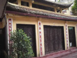 Hình ảnh chụp mặt tiền của một công trình kiến trúc cổ, có thể là một ngôi chùa hoặc đền thờ. Tòa nhà có tường sơn màu vàng, mái ngói cong theo phong cách kiến trúc truyền thống Á Đông. Có nhiều bộ cửa gỗ lớn màu nâu sẫm, một số được chạm khắc hoa văn, và tất cả đều đang đóng. Hai bên các khung cửa có các cột trụ treo câu đối dọc màu đỏ, viết chữ Hán màu trắng. Nội dung các câu đối có thể đọc được như sau:。- Cột bên trái: "南天聖" (Nam Thiên Thánh).。- Cột ở giữa, bên trái cửa lớn: "真儒佛市在湖神" (Chân Nho Phật Thị Tại Hồ Thần).。- Cột ở ngoài cùng bên phải: "古今士子拜師顏" (Cổ Kim Sĩ Tử Bái Sư Nhan).。Phía trước tòa nhà có một bụi cây xanh và các bậc thềm bằng đá màu đỏ dẫn lên trên. Ở góc phải có một tấm biển nhỏ màu trắng nhưng không thể đọc rõ nội dung. Khung cảnh tĩnh lặng, không có hoạt động nào của con người được ghi lại.。（日本語訳）。古い建築物の正面を撮影した写真で、寺院か祠である可能性があります。建物は壁が黄色に塗られ、東アジアの伝統的な建築様式に沿った湾曲した瓦屋根を持っています。濃い茶色の大きな木製の扉が多数あり、そのうちのいくつかは模様が彫刻されており、すべて閉まっています。ドア枠の両側には、白い漢字が書かれた赤い縦長の対聯が掛けられた柱があります。対聯の内容は以下のように読み取れます：。- 左側の柱：「南天聖」（ナム・ティエン・タイン）。- 中央、大きな扉の左側の柱：「真儒佛市在湖神」（チャン・ニョー・ファット・ティ・タイ・ホー・タン）。- 一番右側の柱：「古今士子拜師顏」（コー・キム・シー・トゥ・バイ・スー・ニャン）。建物の前には緑の茂みと、上へと続く赤い石の階段があります。右隅には小さな白い看板がありますが、内容ははっきりと読み取れません。静かな光景で、人の活動は記録されていません。